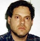 A man with short, dark hair and a stern expression, wearing a plaid shirt against a white background.