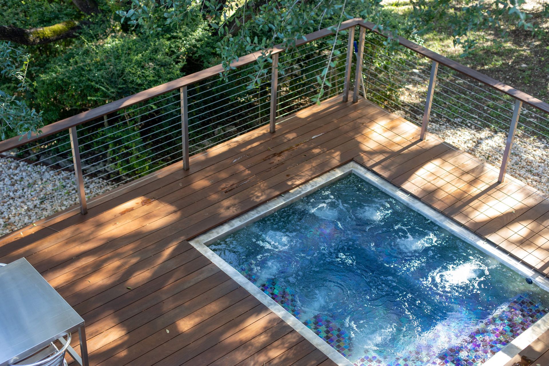 Wooden deck stainless steel hot tub surrounded by metal railings and lush greenery.