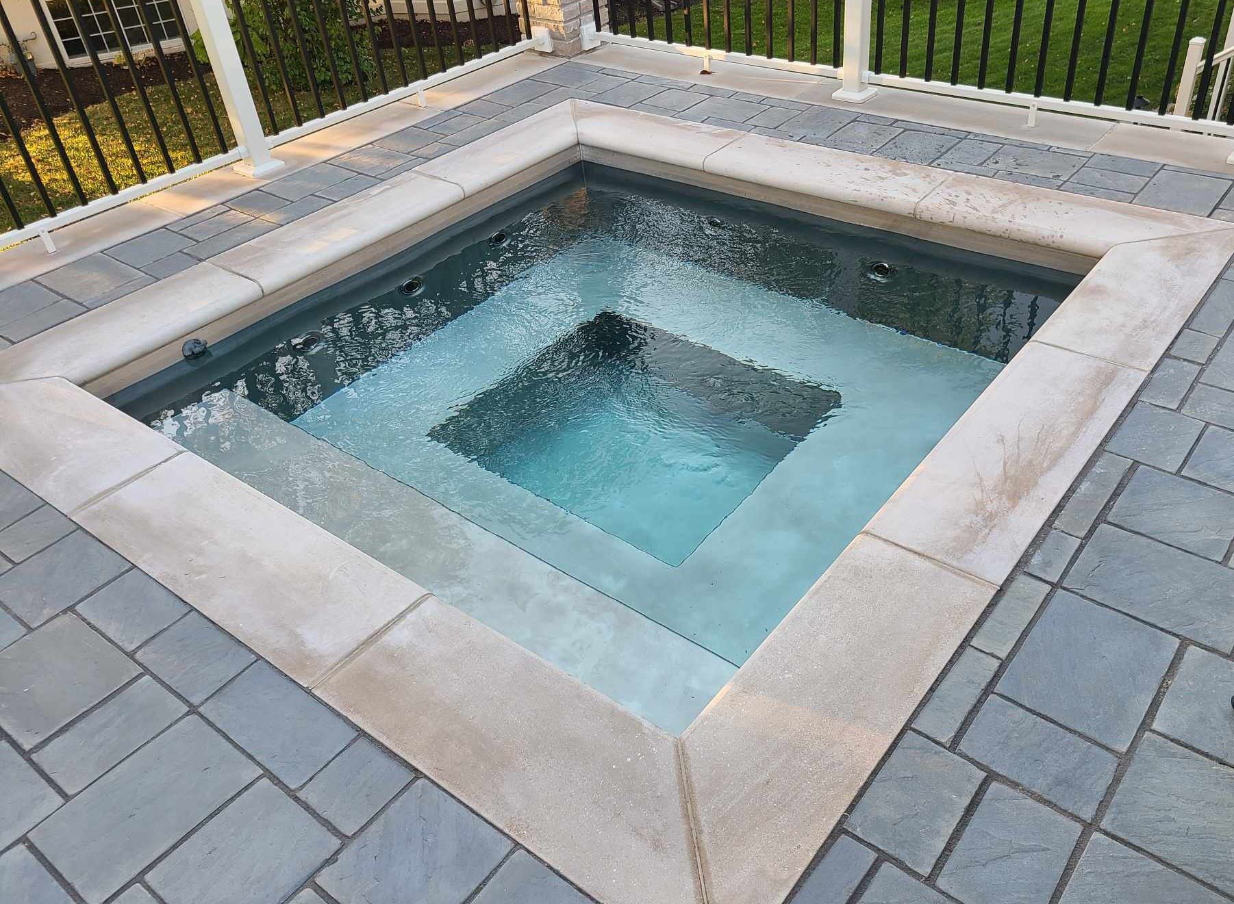 Square outdoor hot tub with tiered steps, surrounded by gray paving stones.