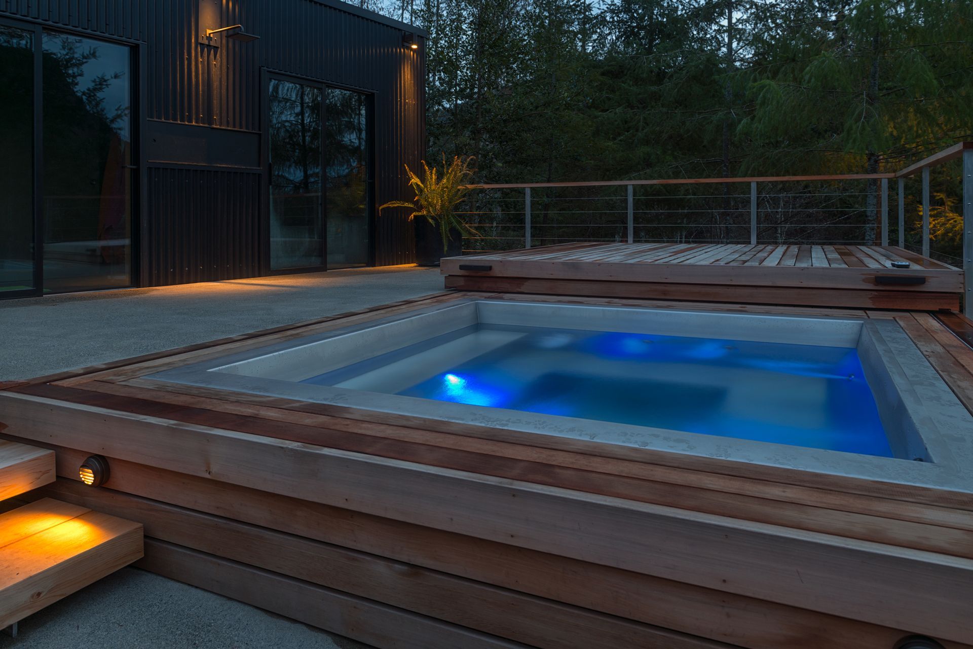 Hot tub with blue lights, surrounded by wooden decking, near a modern dark building.