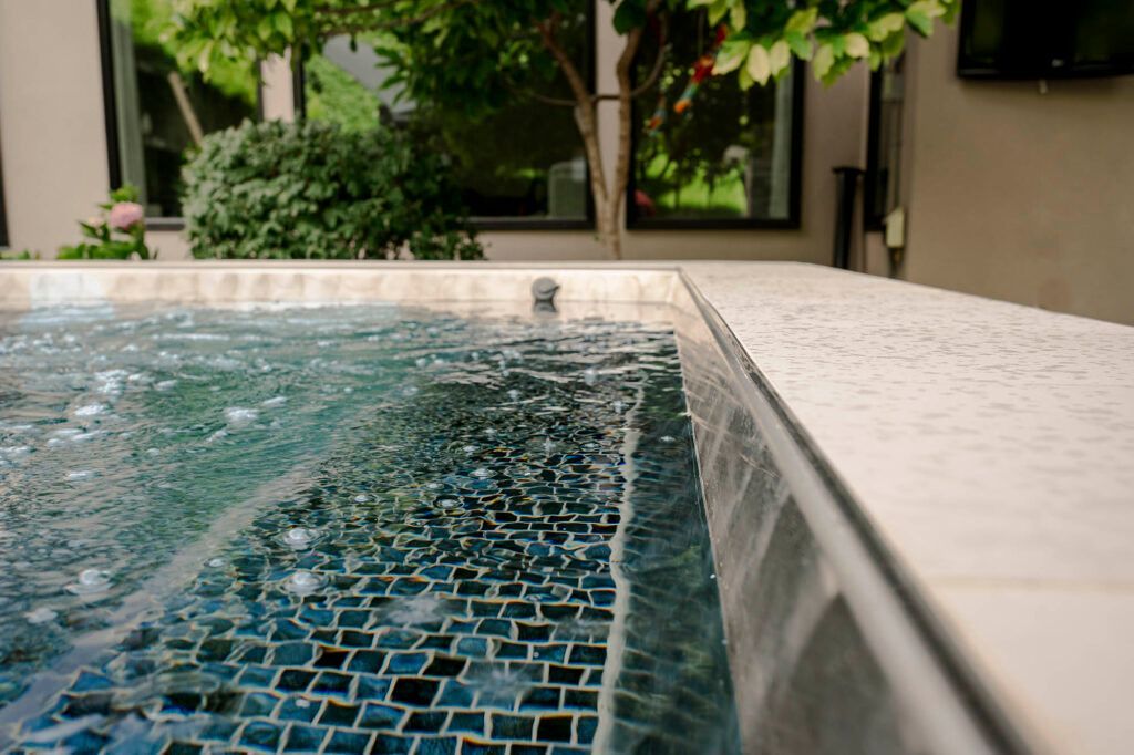 Spa with blue mosaic tiles and bubbling water, stone coping, and greenery in the background.