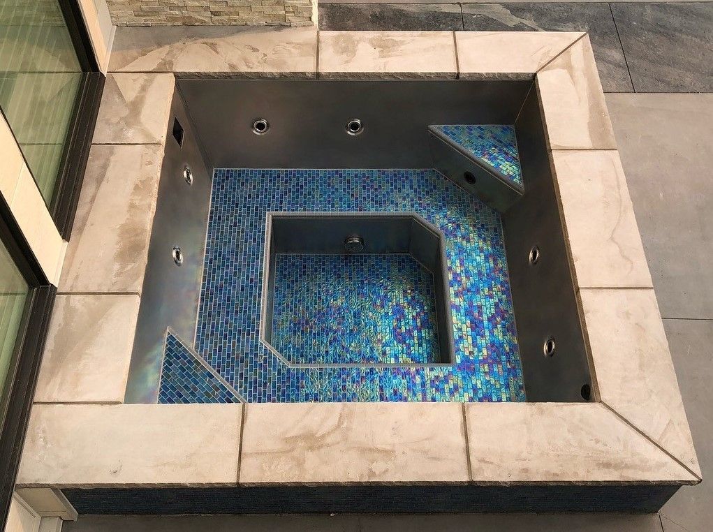 Square blue-tiled hot tub built into a stone patio, surrounded by gray stone.