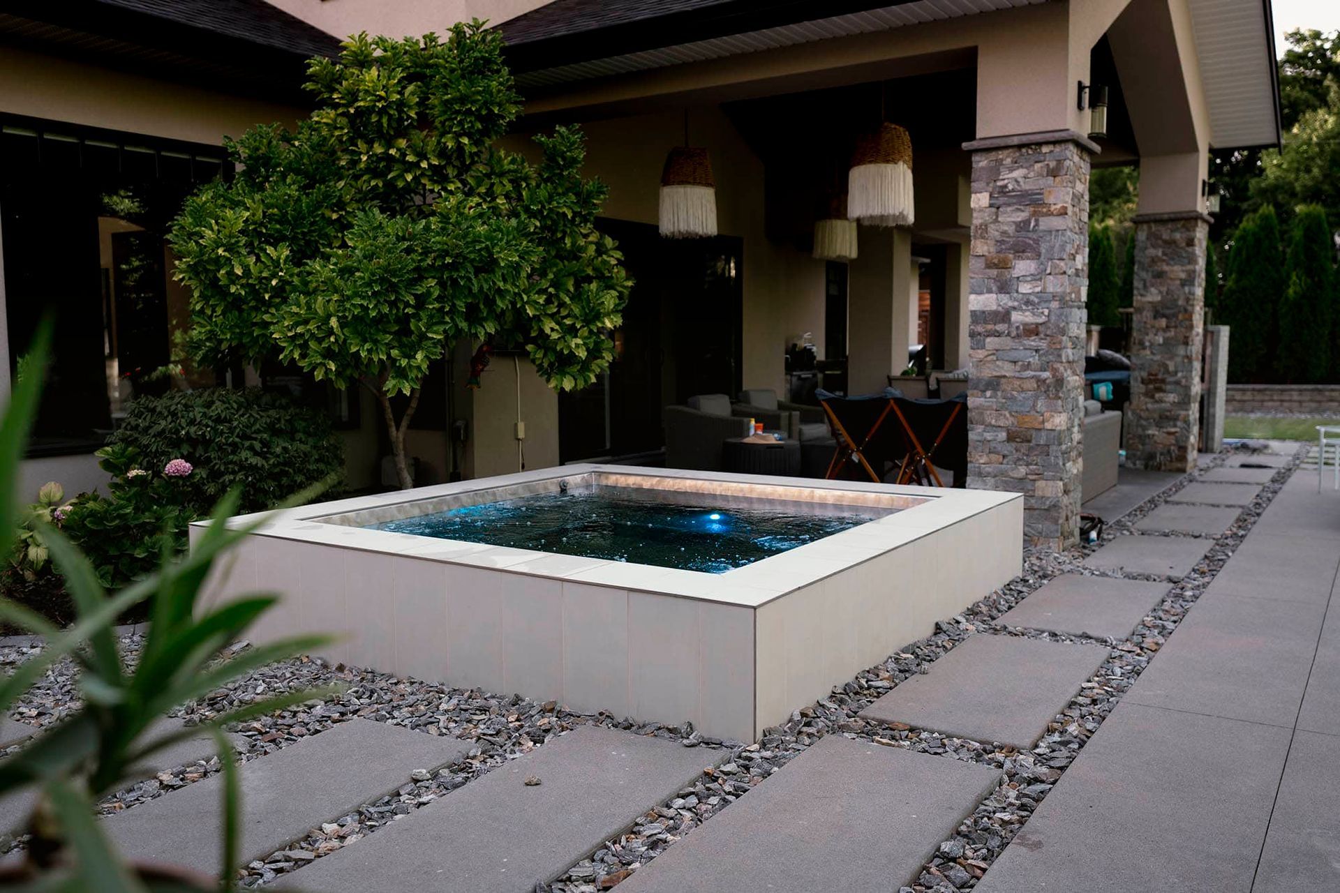 A square hot tub with blue water on a stone patio, adjacent to a house with a covered porch.