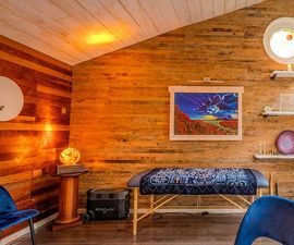 A therapy room with wood-paneled walls, a massage table with a patterned blue cover, warm lighting, and a framed painting.