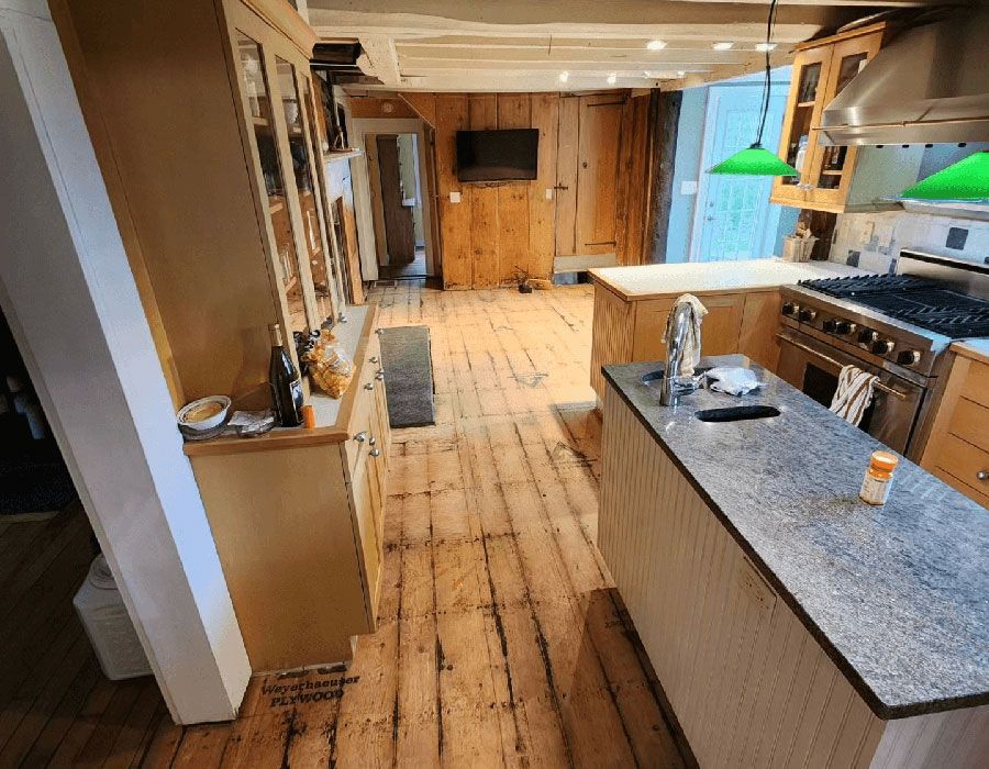 A bright kitchen with light wooden floors, a granite-topped island, wooden cabinetry, and a stainless steel stove.