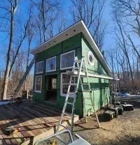 Green tiny house under construction with ladder outside in a wooded yard