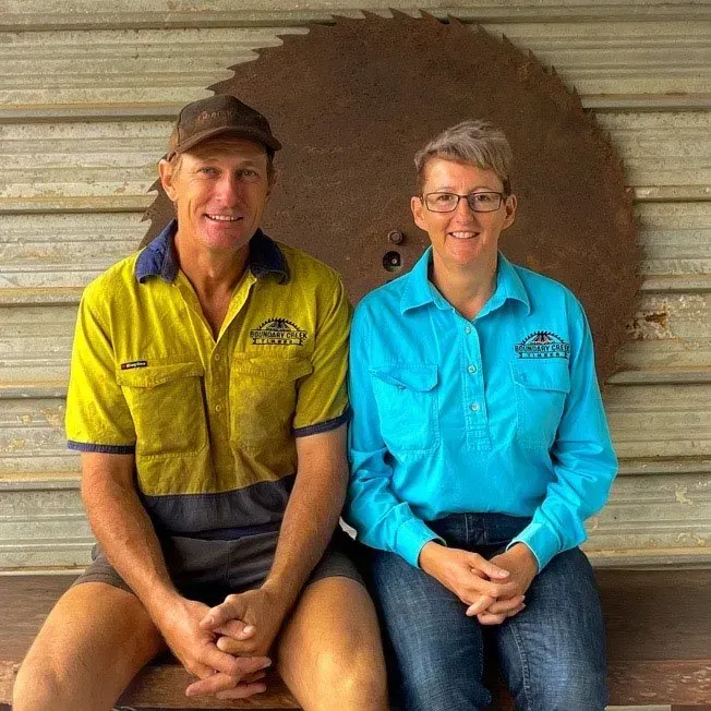 Two people in work clothes sit on a bench; a rusty saw blade hangs behind them — Boundary Creek Timber in Wallaville, QLD