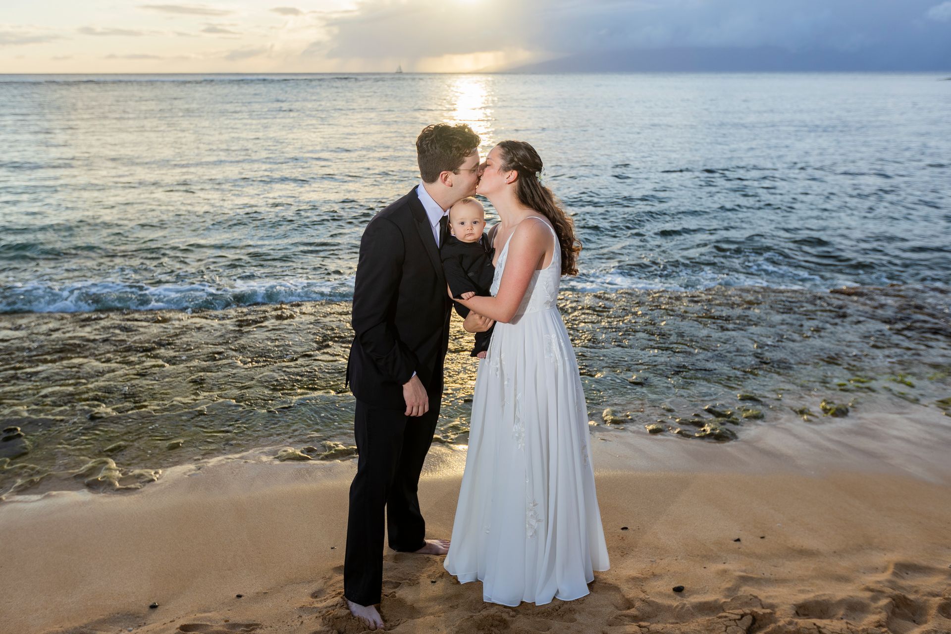 Couple kissing on beach, holding baby at sunset.