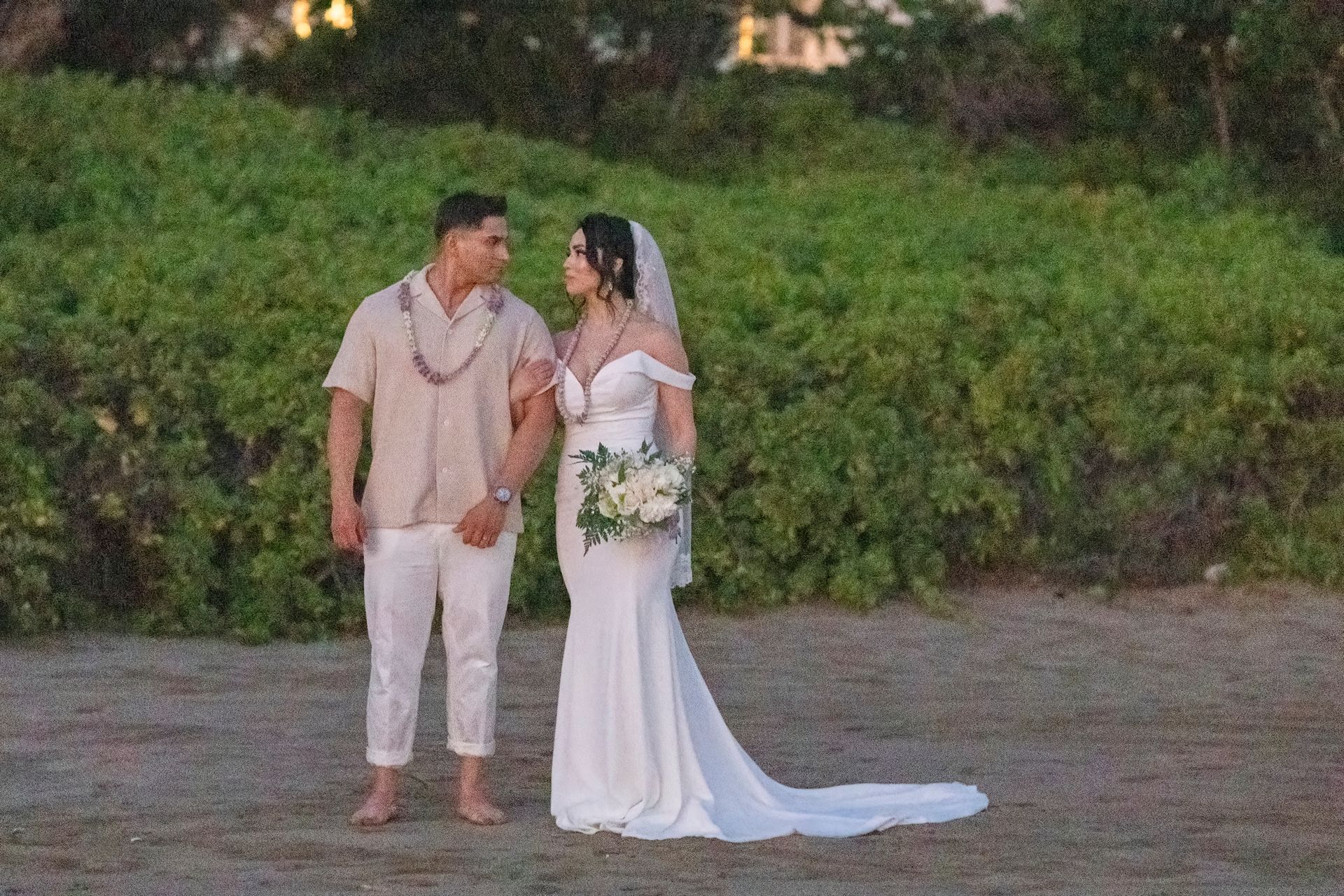Newlyweds on a beach, gazing at each other. The bride in white gown, groom in beige shirt and white pants.