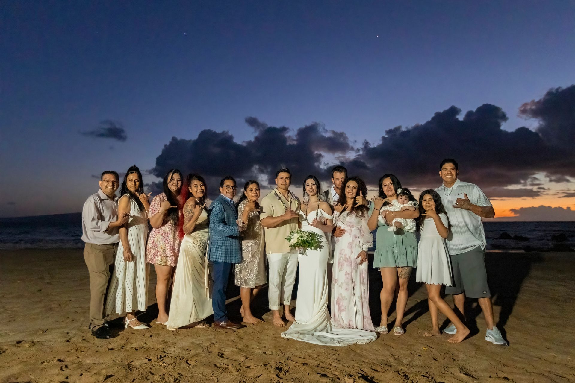 Group of people on beach at sunset, bride and groom in front, others smiling, dark sky.