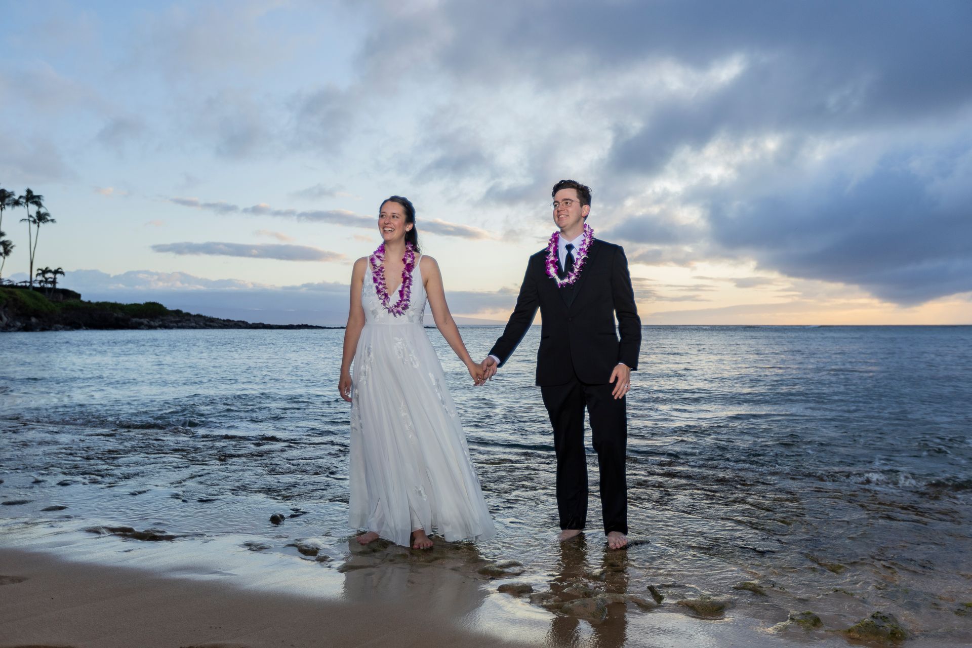 Bride and groom holding hands, standing in the ocean at sunset, wearing leis.