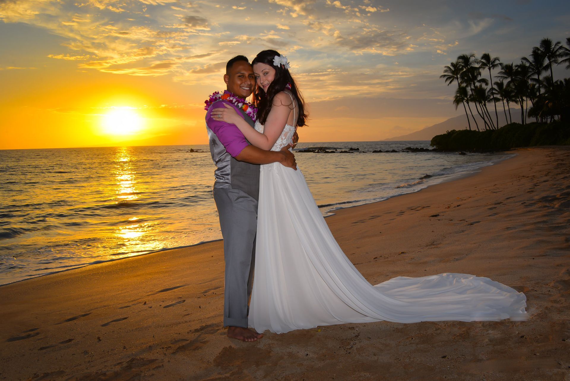 Couple hugging on a beach at sunset. Bride in white dress, groom in purple shirt, sun over the ocean.
