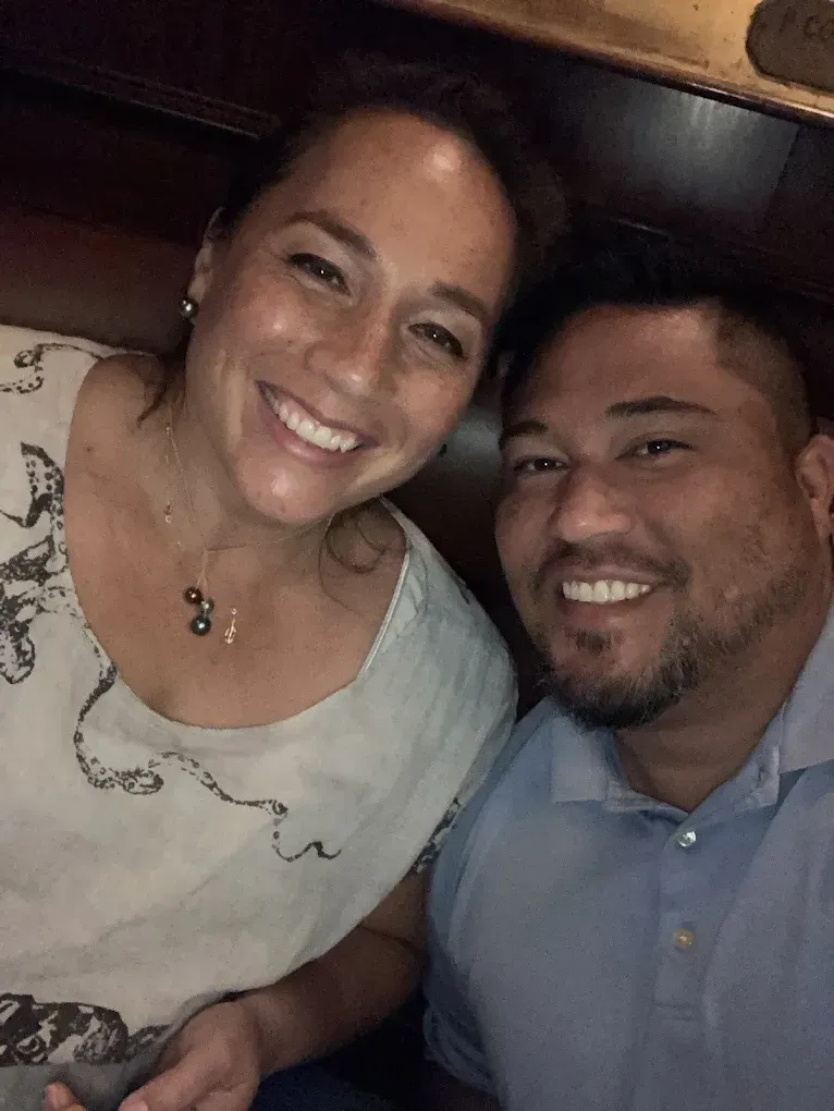 Smiling woman and man posing together, lit from below. Woman in patterned top, man in blue shirt. Dark background.