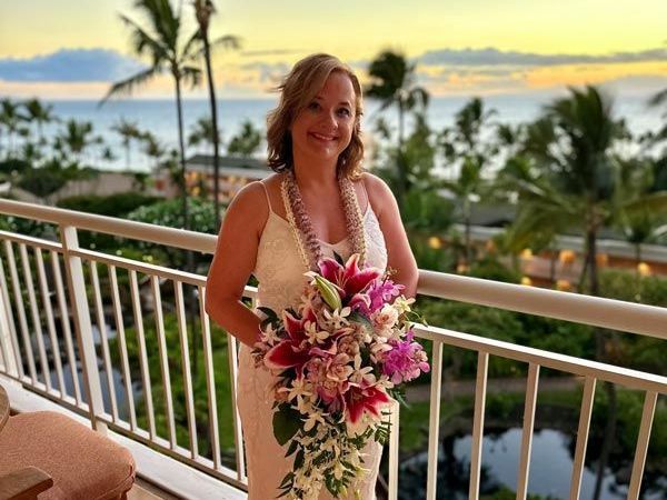 Woman on a balcony with a bouquet of flowers and leis, smiling with an ocean sunset in the background.