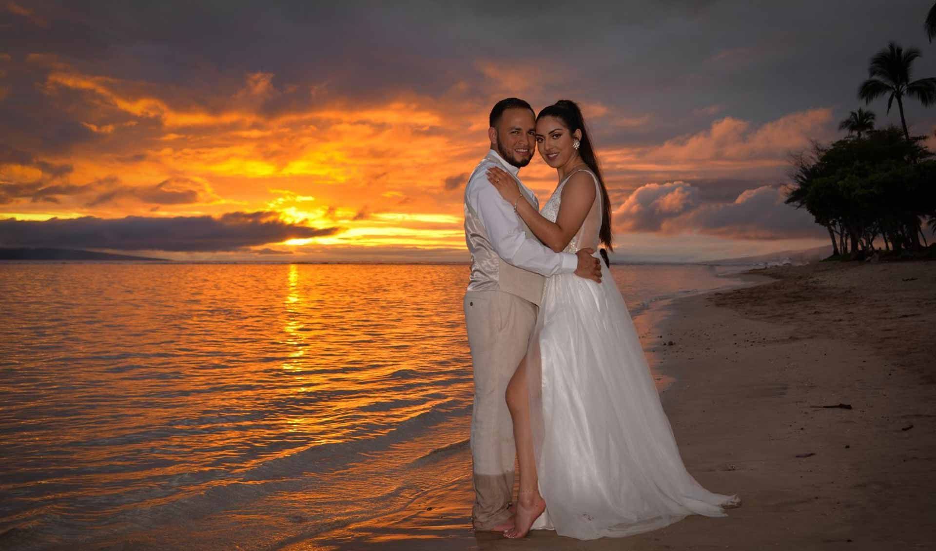 Bride and groom embrace on a beach as the sunset paints the sky orange and the water.