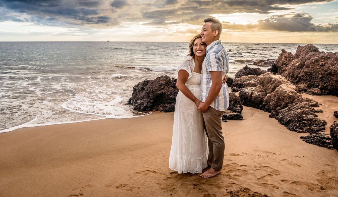 Couple embraces on a sandy beach at sunset. The woman wears a white dress, the man a patterned shirt.