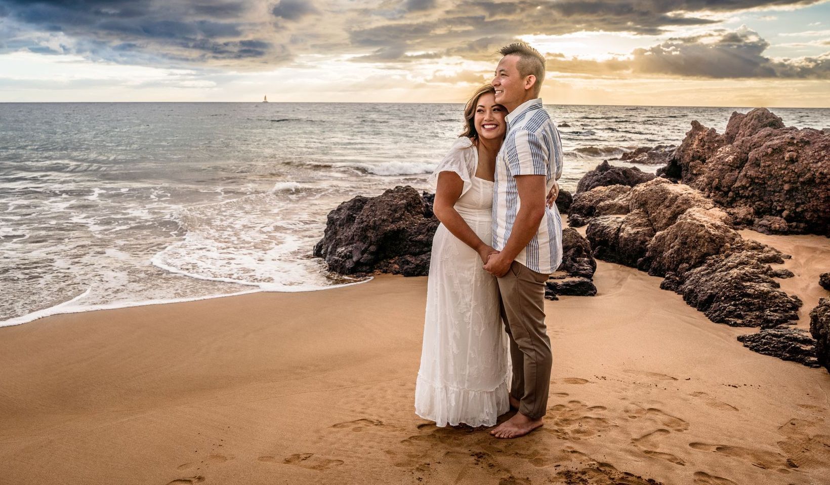 Couple embracing on a beach. Woman in white dress, man in floral shirt. Sunset colors, ocean in background.