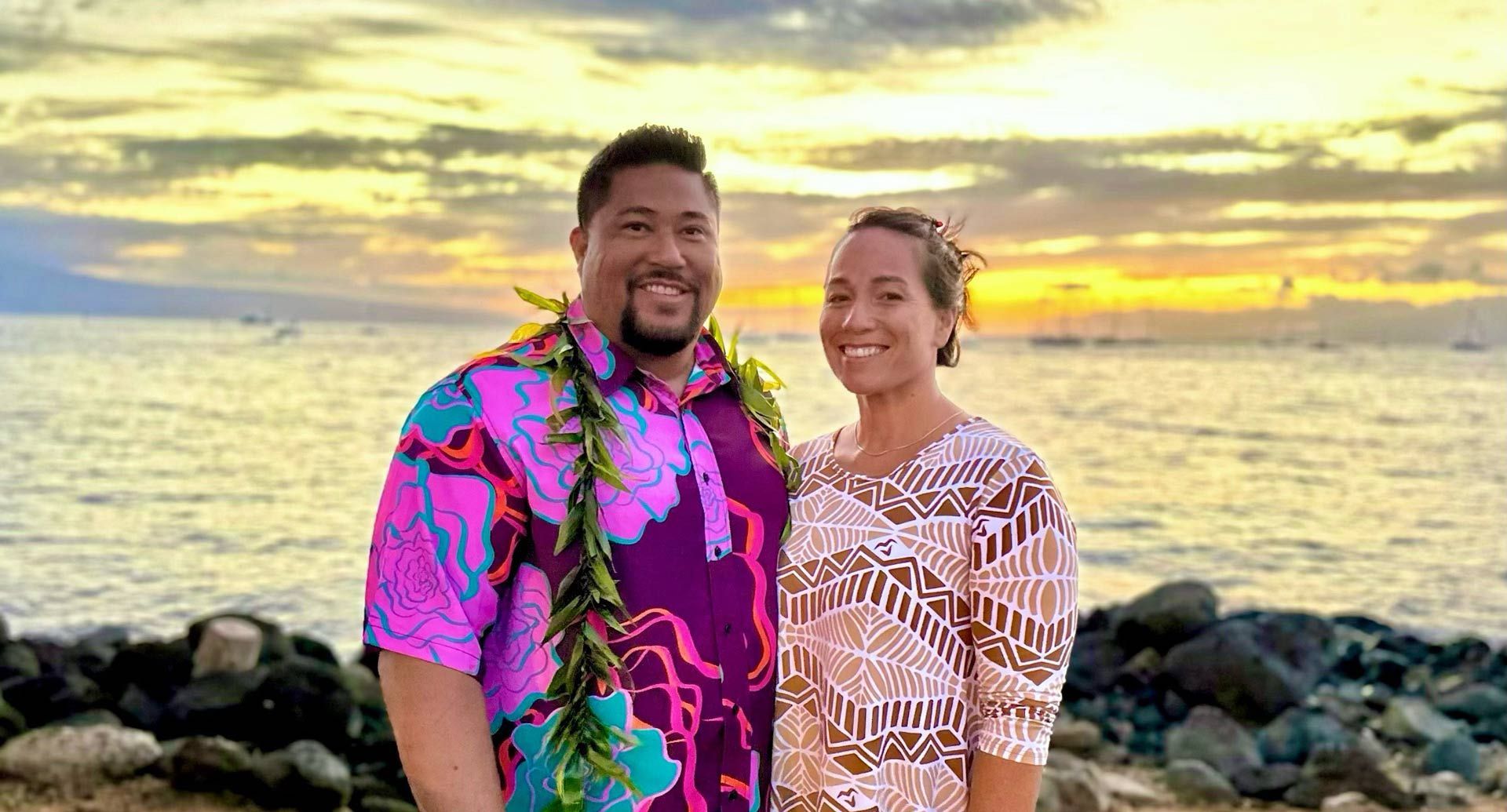 Couple poses for photo on beach at sunset. Man wears floral shirt, woman a patterned top.
