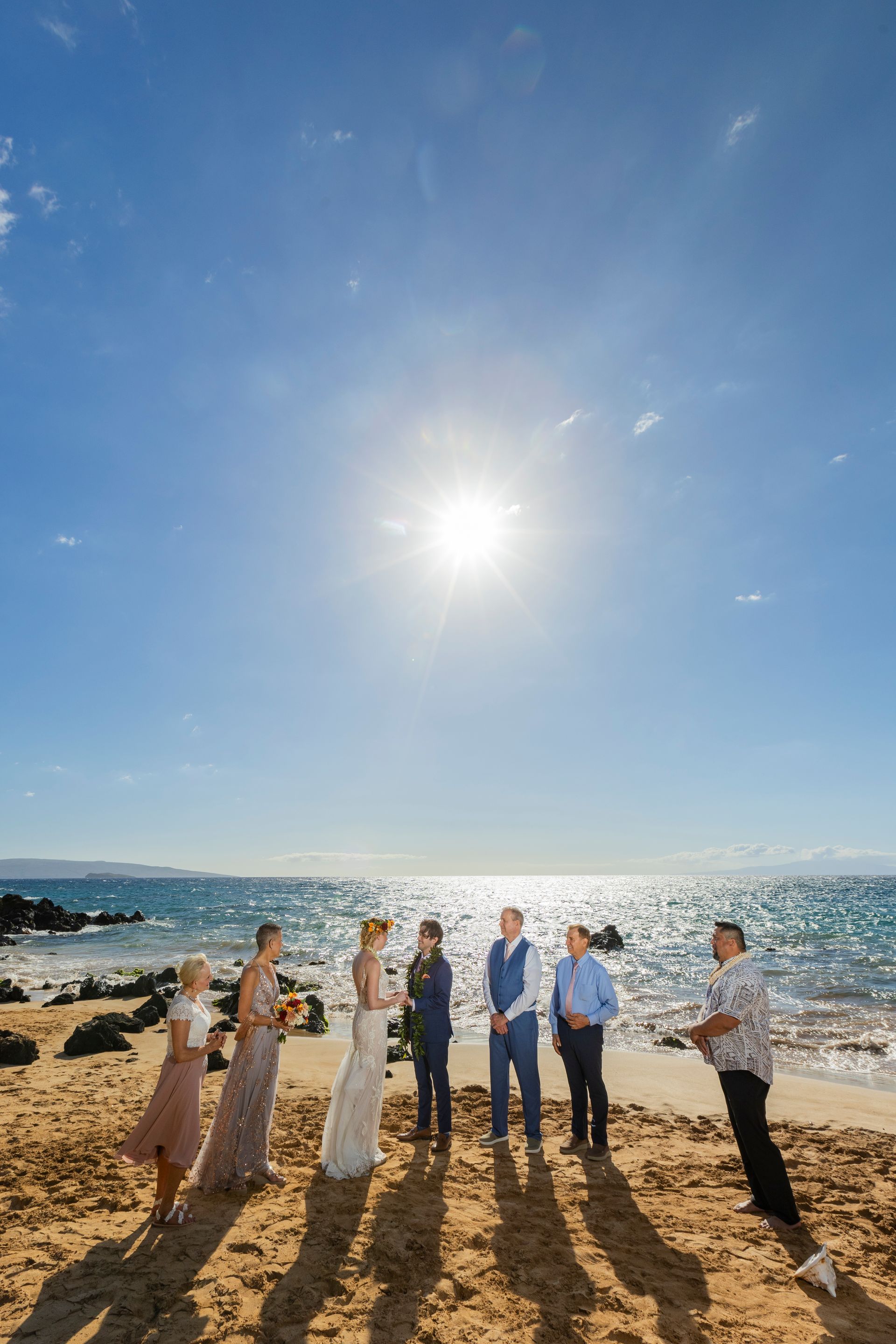 Wedding ceremony on a sunny beach with couple and guests; ocean and blue sky in the background.