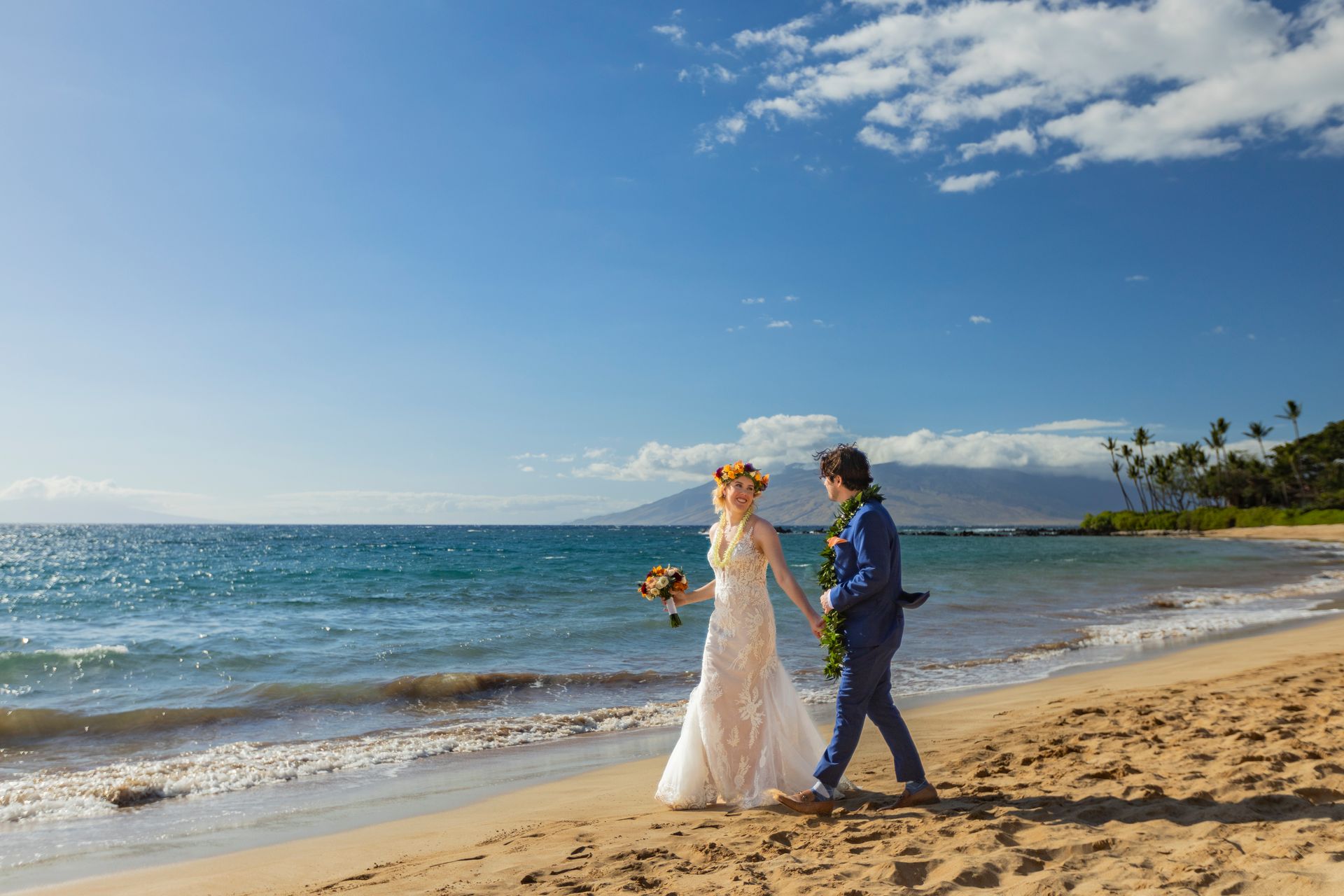 Bride and groom walk hand-in-hand on a sunny beach. Blue ocean, sky, and sand surround them.