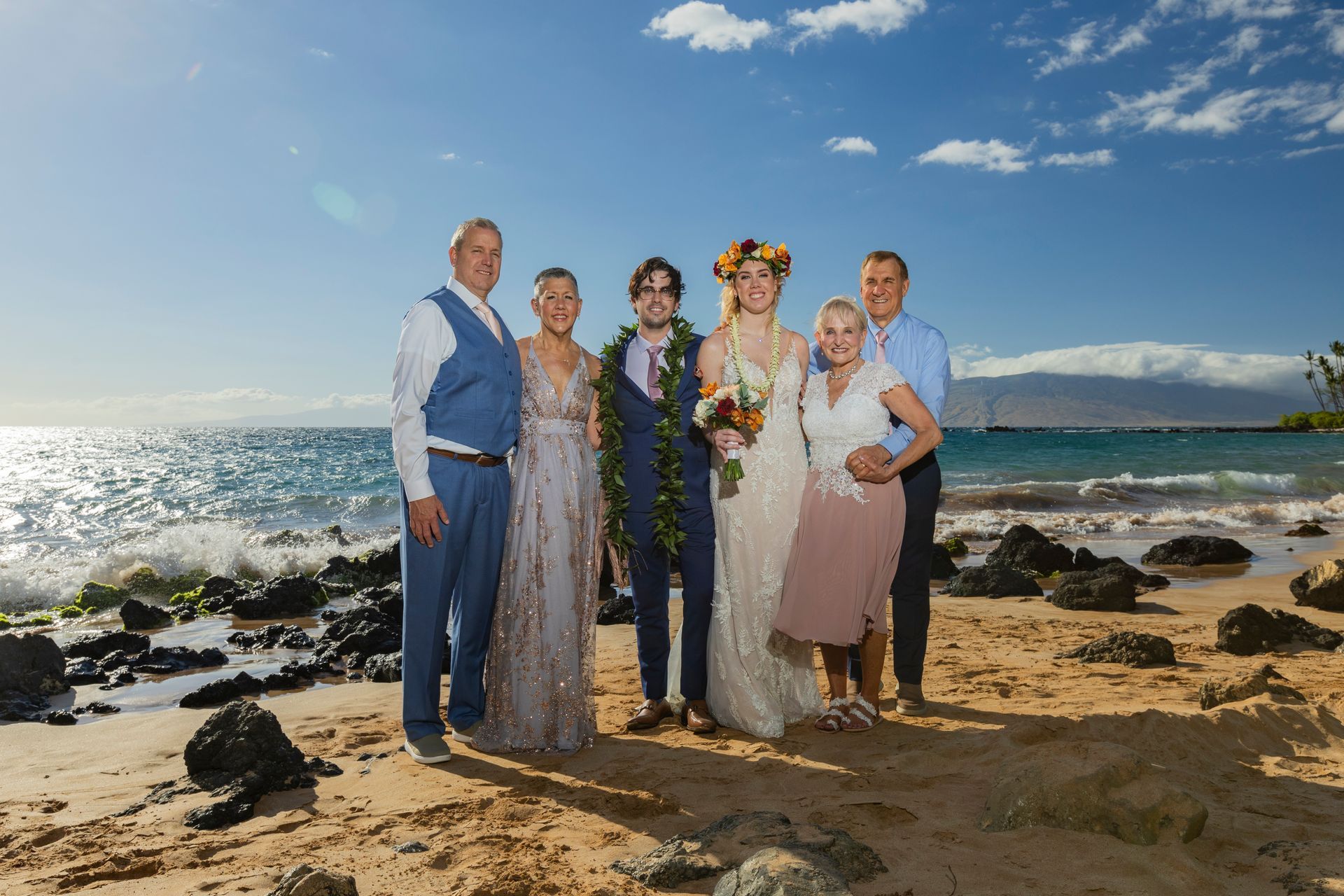 Family wedding portrait on a sunny beach; couple in wedding attire with parents and sibling.