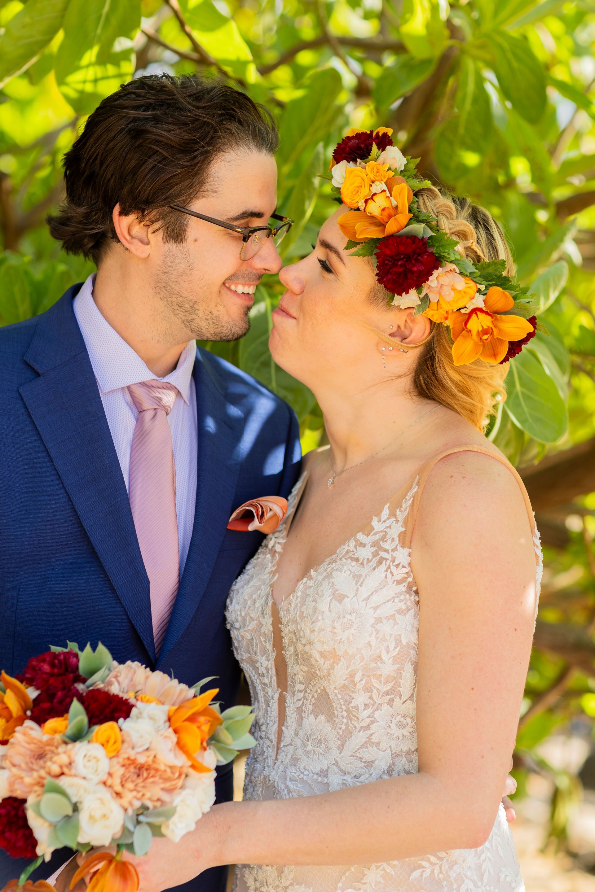 Wedding couple, woman in floral crown and lace dress, man in blue suit and glasses, smiling and touching noses.