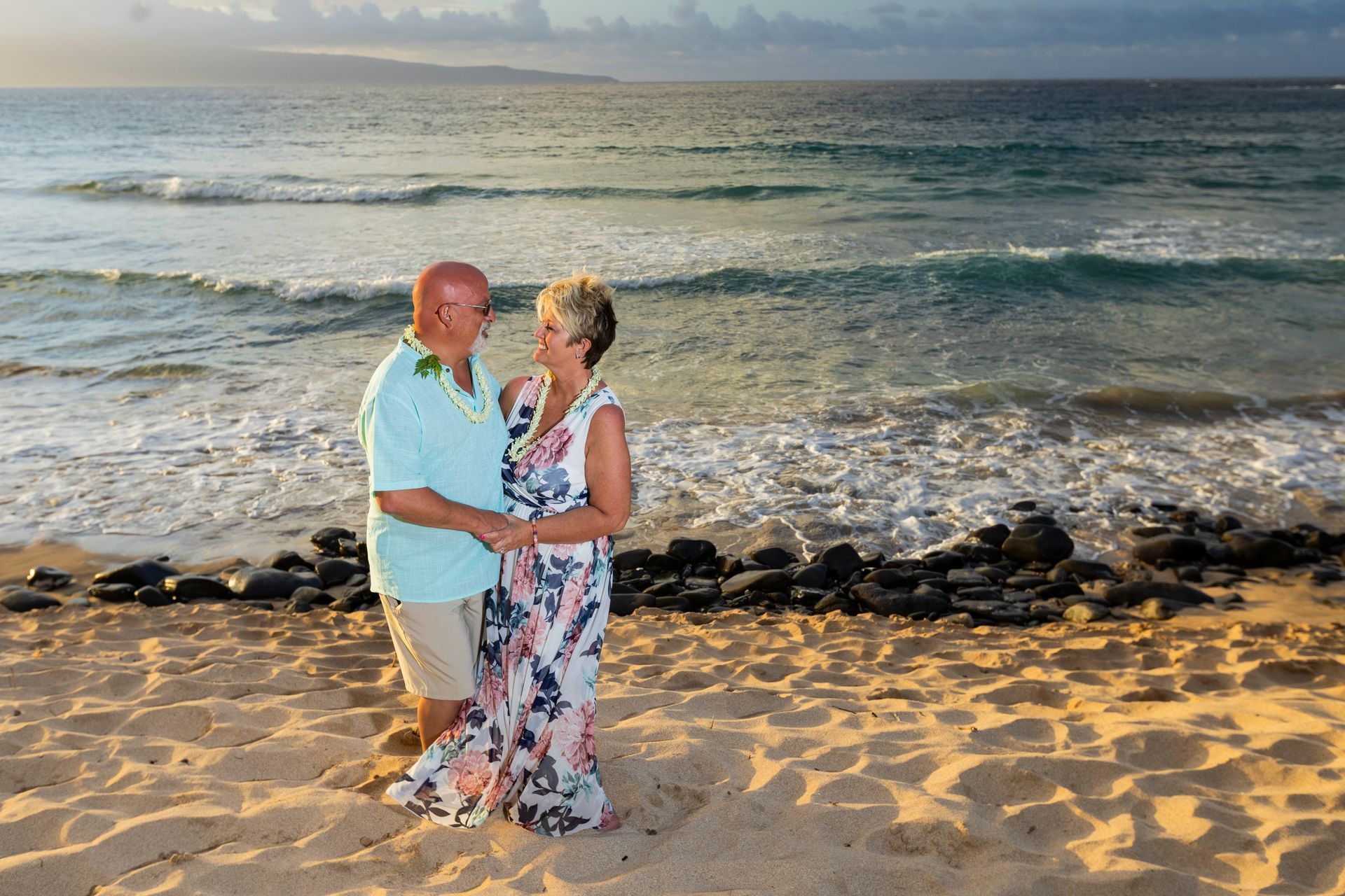 Couple holding hands on a beach, ocean waves and sunset in the background.
