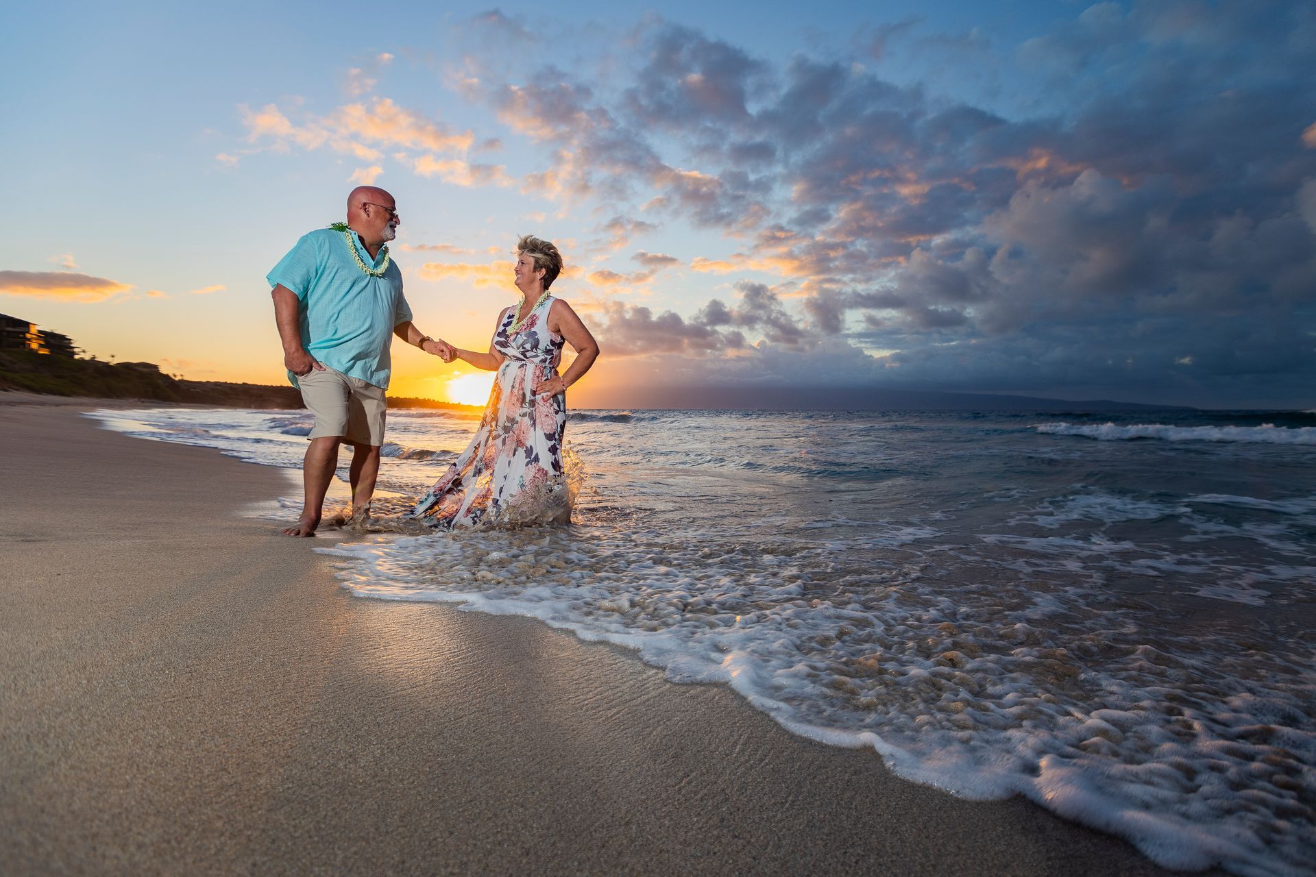 Couple on a beach at sunset, holding hands, enjoying the water. Vibrant sky, light sand, and ocean waves.