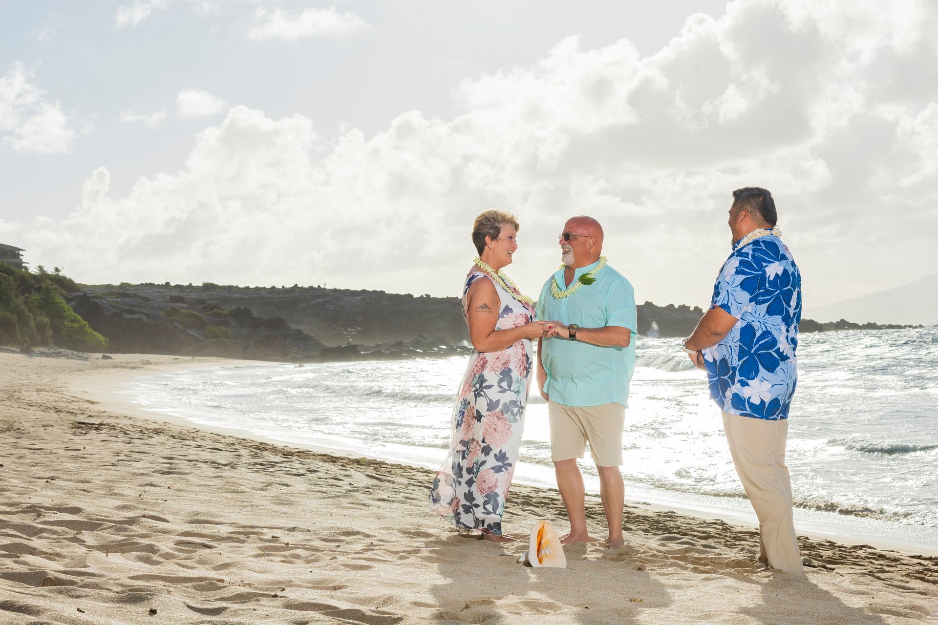 Couple exchanging vows on a beach; officiant in floral shirt, sunny day, ocean waves.