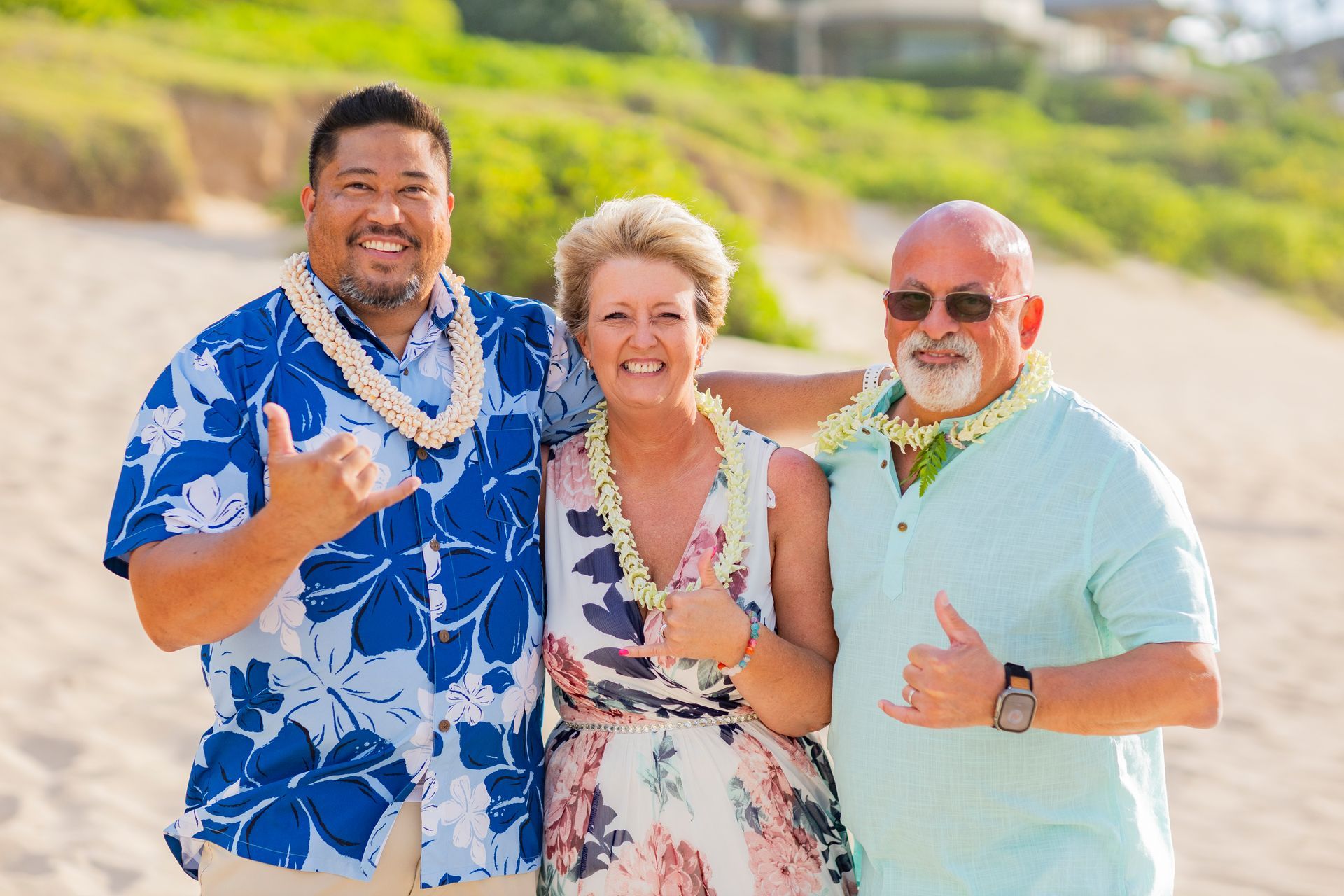 Three people smiling on a beach, wearing leis and casual clothes, giving shaka signs.