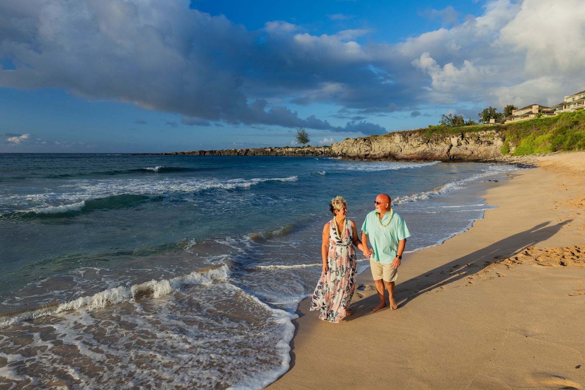 Elderly couple walks along a beach at sunset. The man wears a light blue shirt and shorts, the woman a patterned dress.