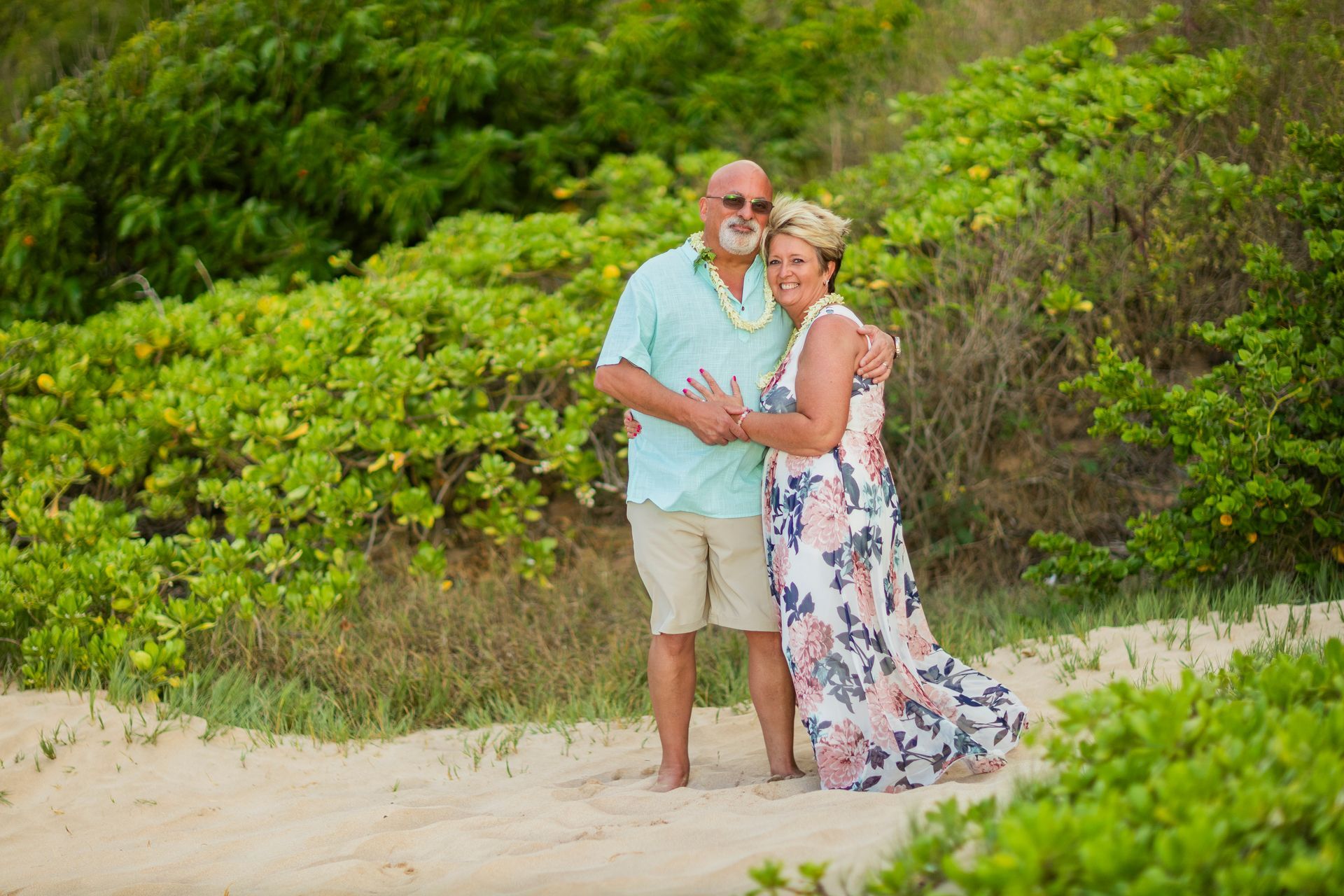Couple hugging on a sandy beach; woman in floral dress, man in teal shirt. Lush green foliage in background.
