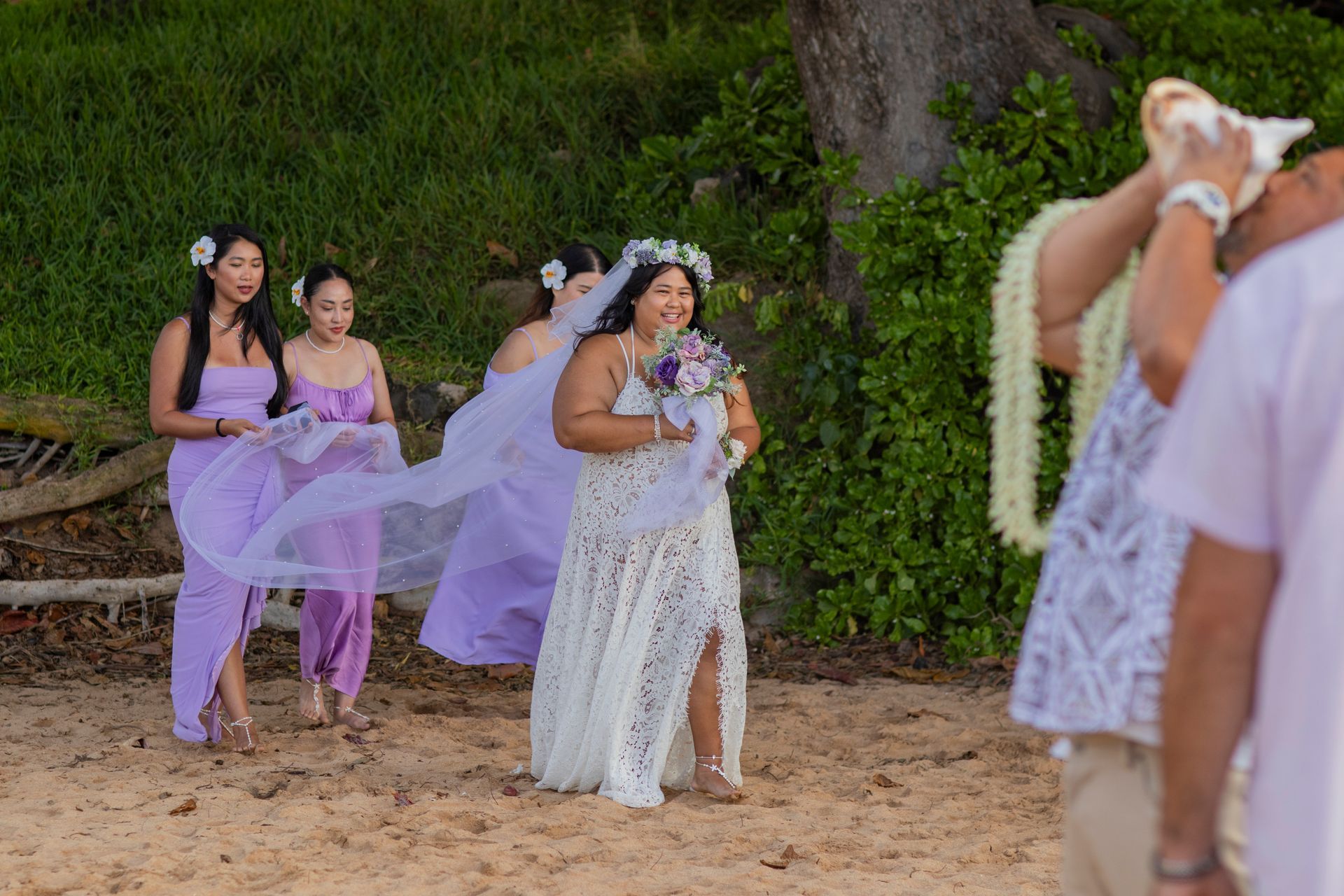 Bride and bridesmaids walk on a beach. Bride wears a lace dress, and bridesmaids are in lavender. A person blows a conch shell.