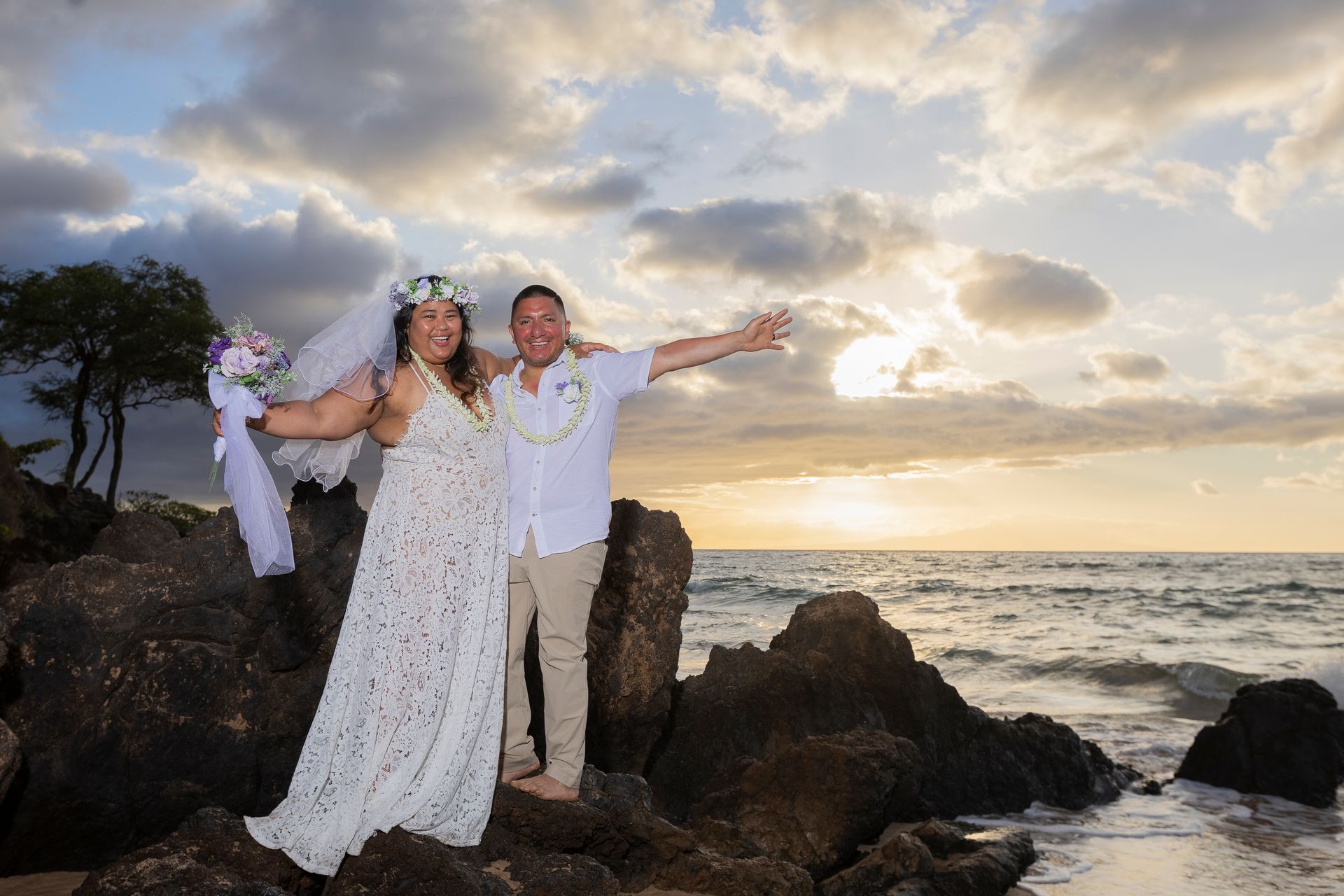 Newlyweds on rocky beach at sunset; woman in white dress and floral crown, man in white shirt with lei, arms outstretched.