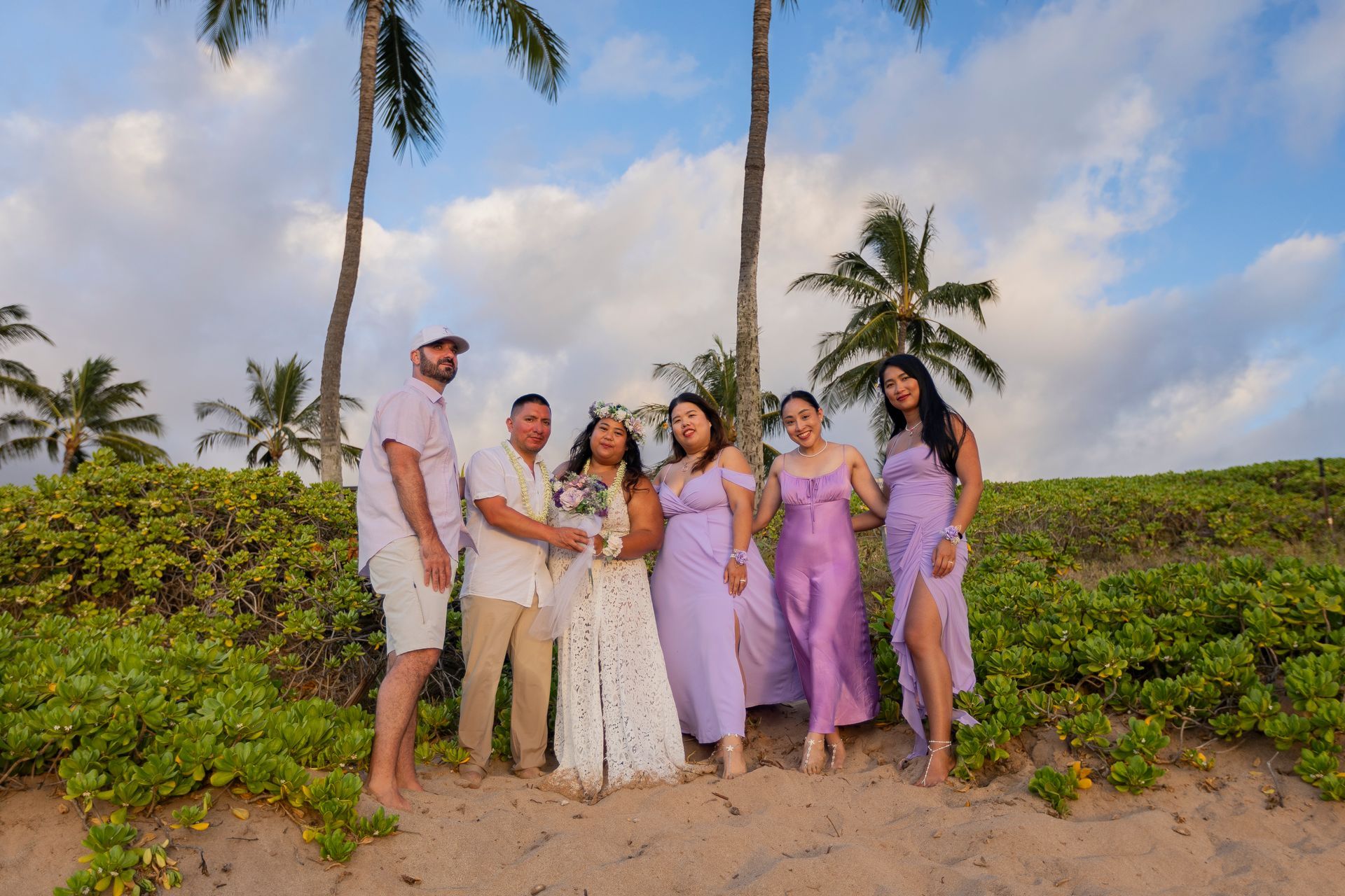Group of people smiling on a beach, wearing light-colored outfits, under palm trees.