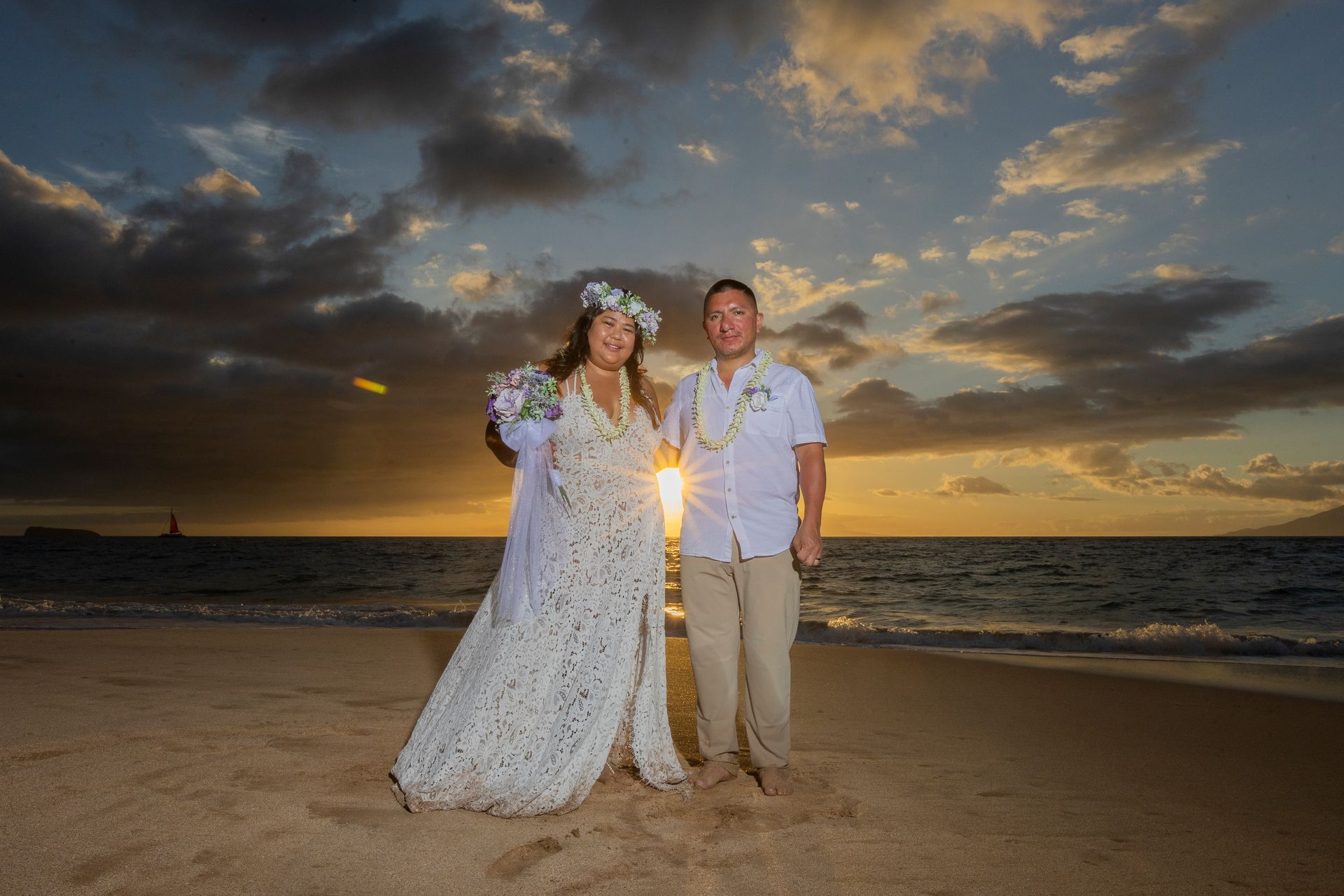 Bride and groom pose on a beach at sunset. The woman holds a bouquet; the man smiles, wearing a lei.