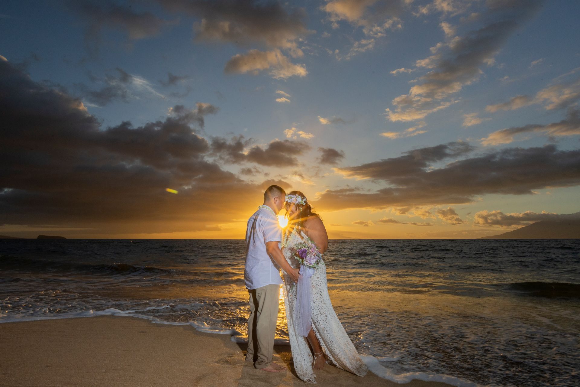 Couple on beach at sunset, holding each other. Bride wears dress, groom, shirt and pants. Ocean in background, clouds.