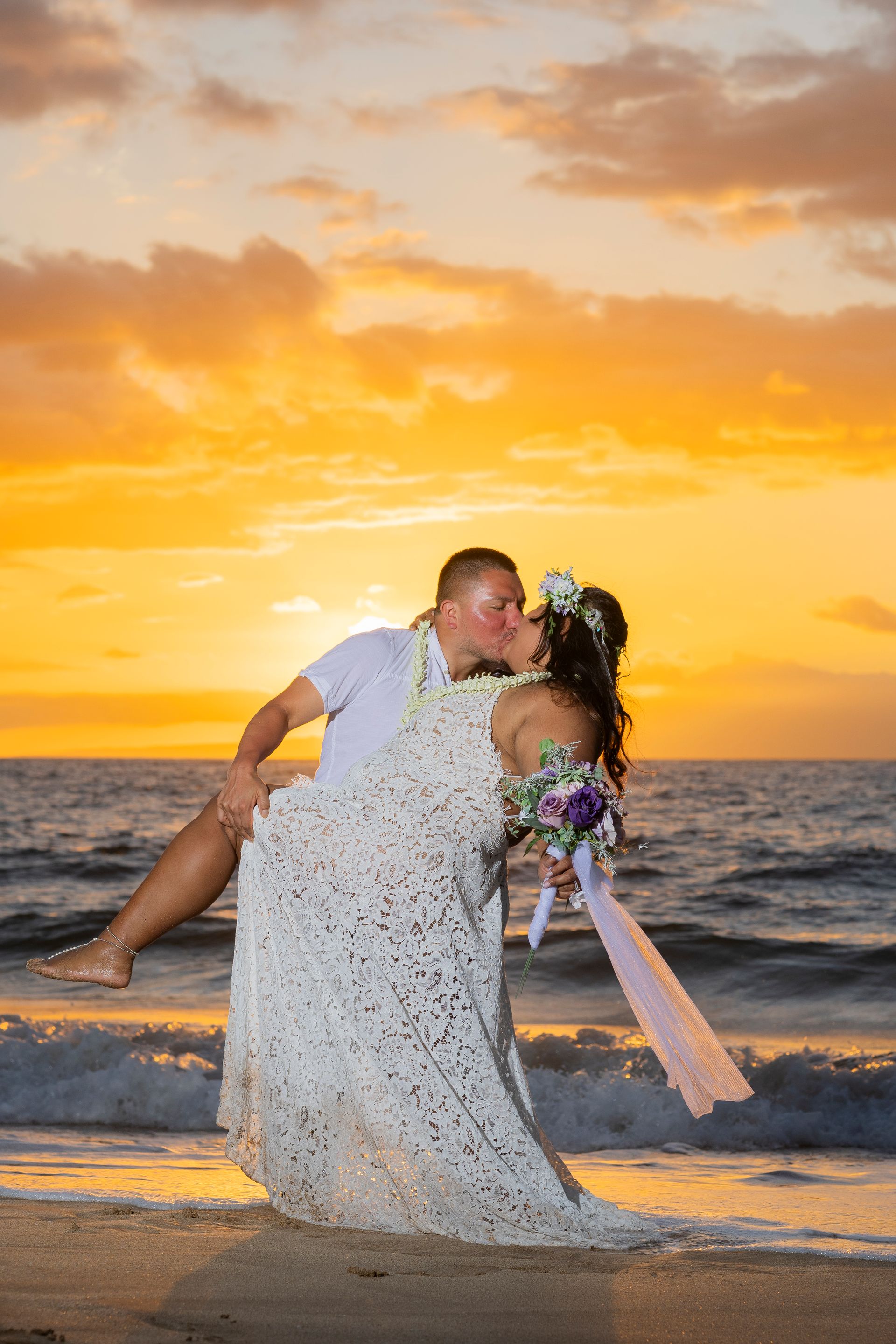 Groom lifts bride, kissing, at sunset on a beach. They're in wedding attire with a colorful sky.