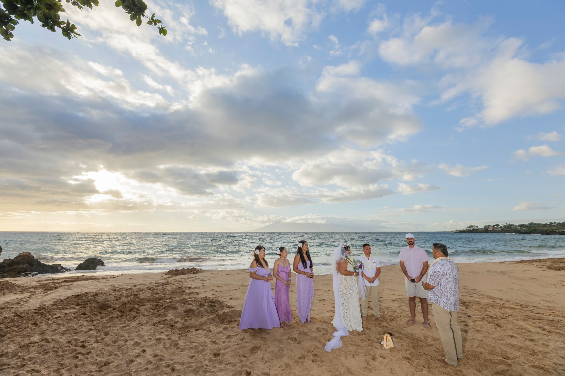 Wedding on beach with bride, bridesmaids, and groom under cloudy sky.