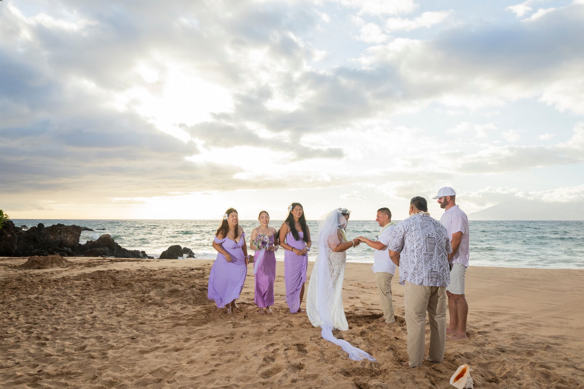 Bride and groom exchange vows on a beach, bridesmaids in purple, groomsmen in light colors; ocean and cloudy sky.