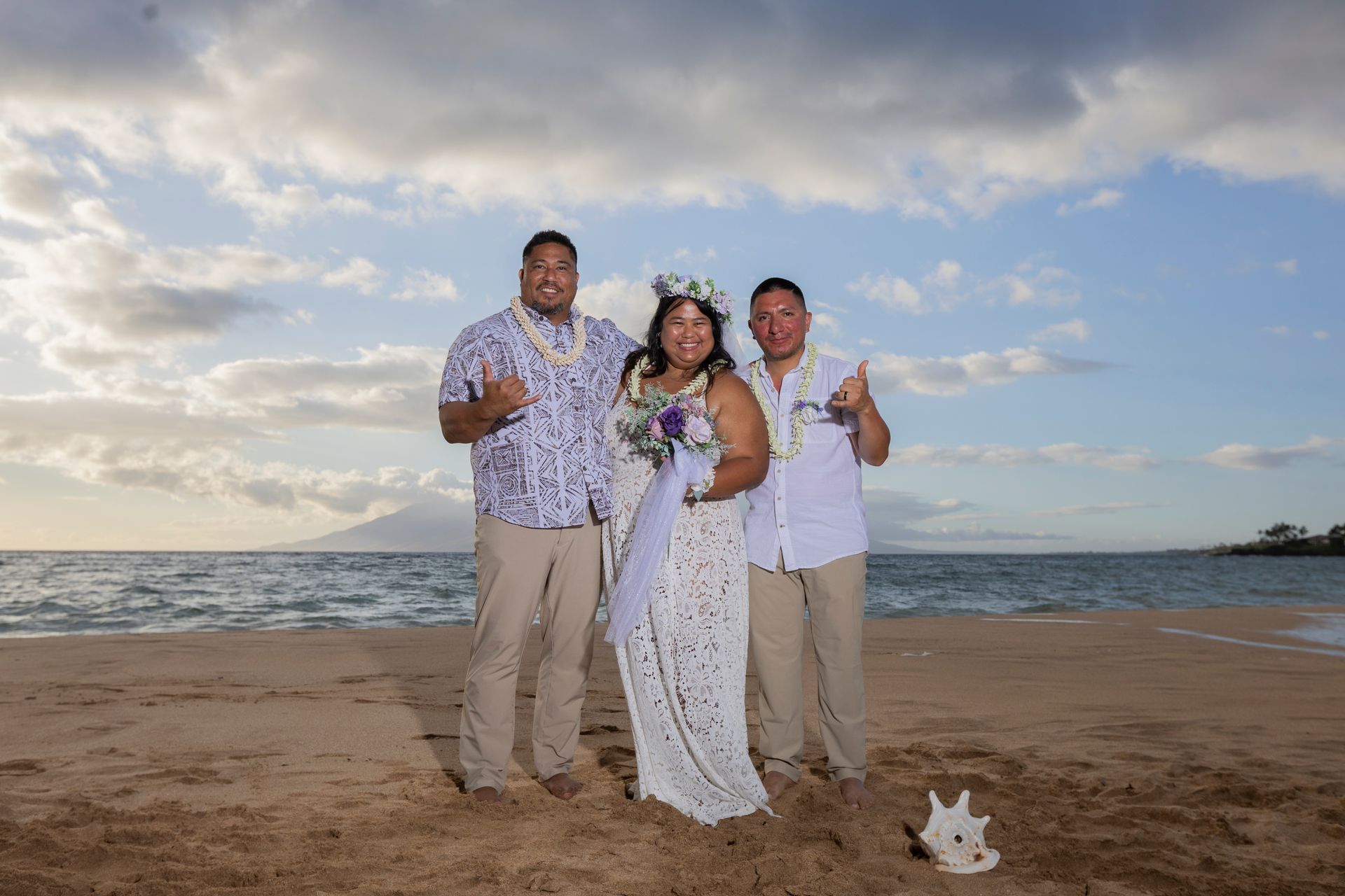 Wedding couple on a beach, flanked by two men, all giving shaka signs. Ocean and sky background.