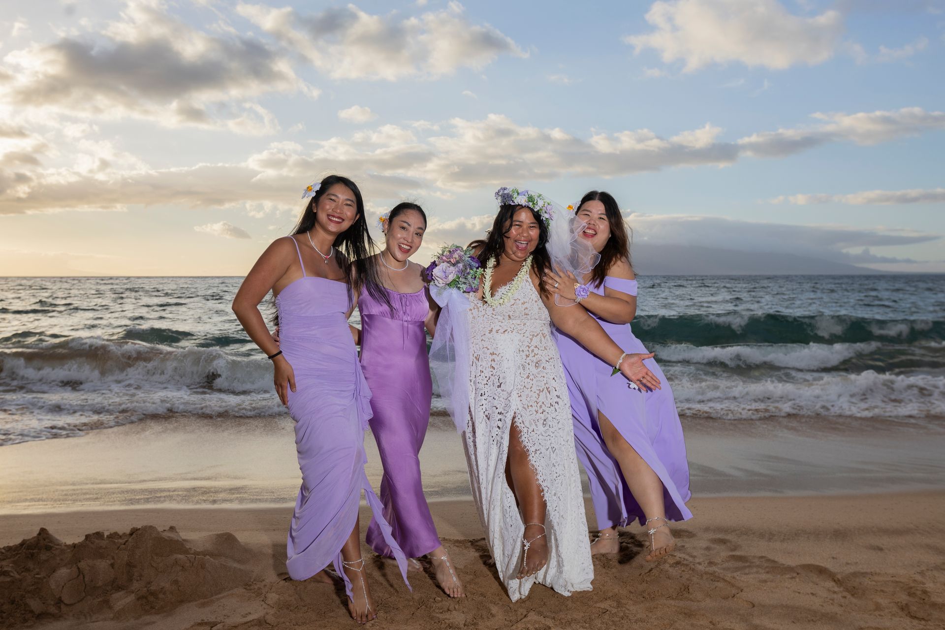 Bride in white dress with bridesmaids in lavender dresses on a beach at sunset.