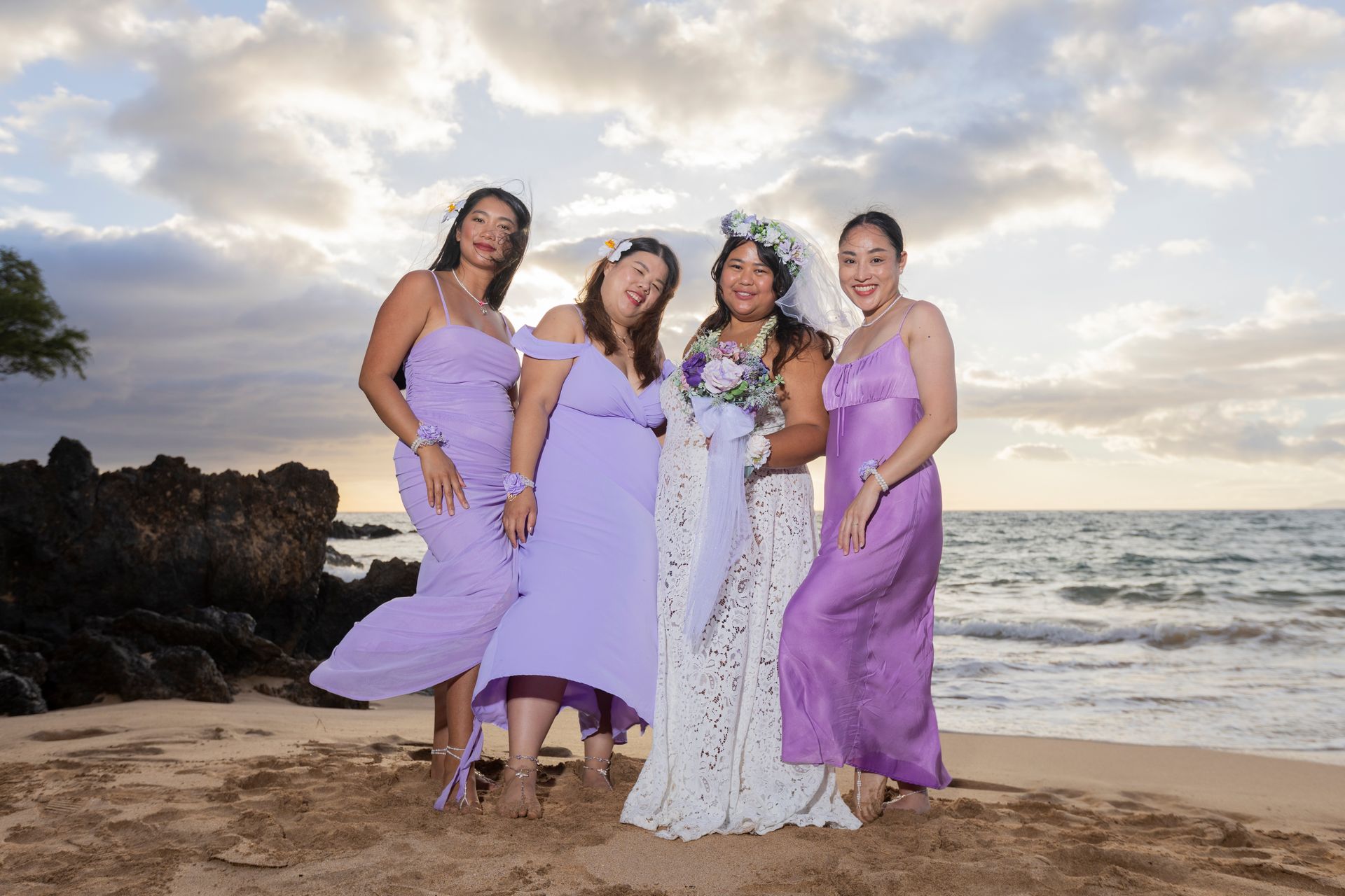 Bride and bridesmaids on beach in lavender dresses at sunset.
