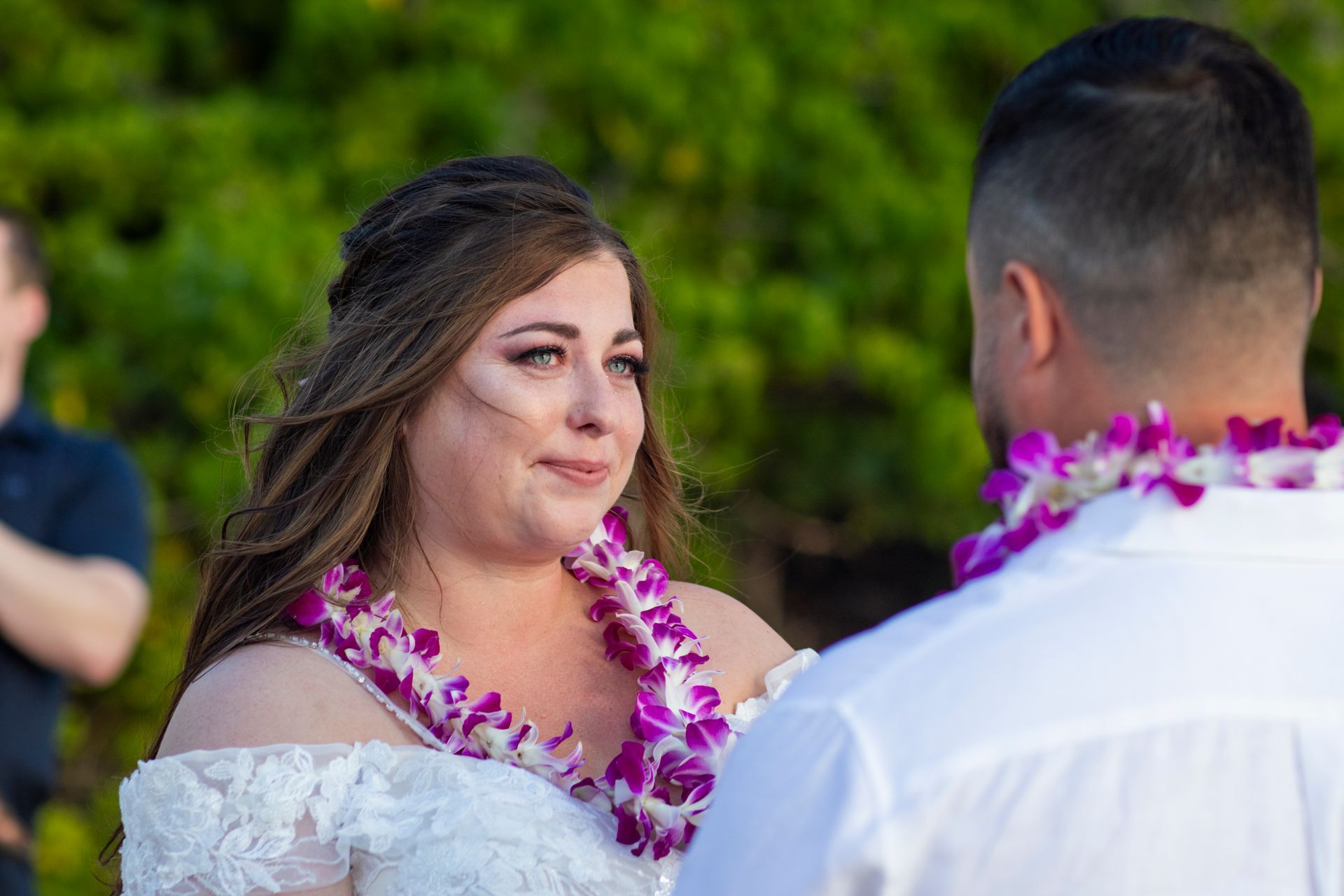 Bride with a lei looks at groom, both in white, during an outdoor wedding.