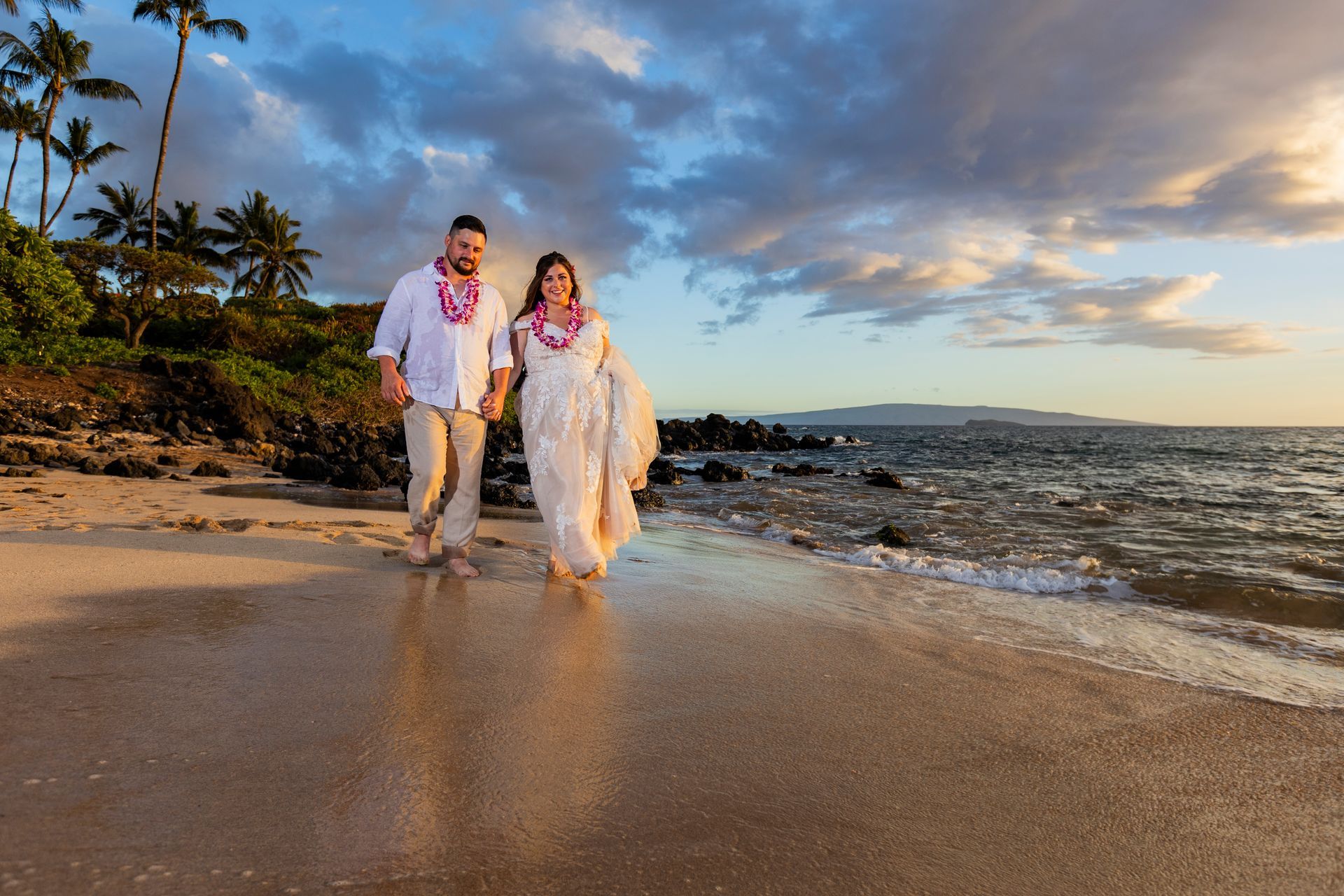 Couple walks barefoot on a beach, holding hands. Ocean waves, sunset light, leis.