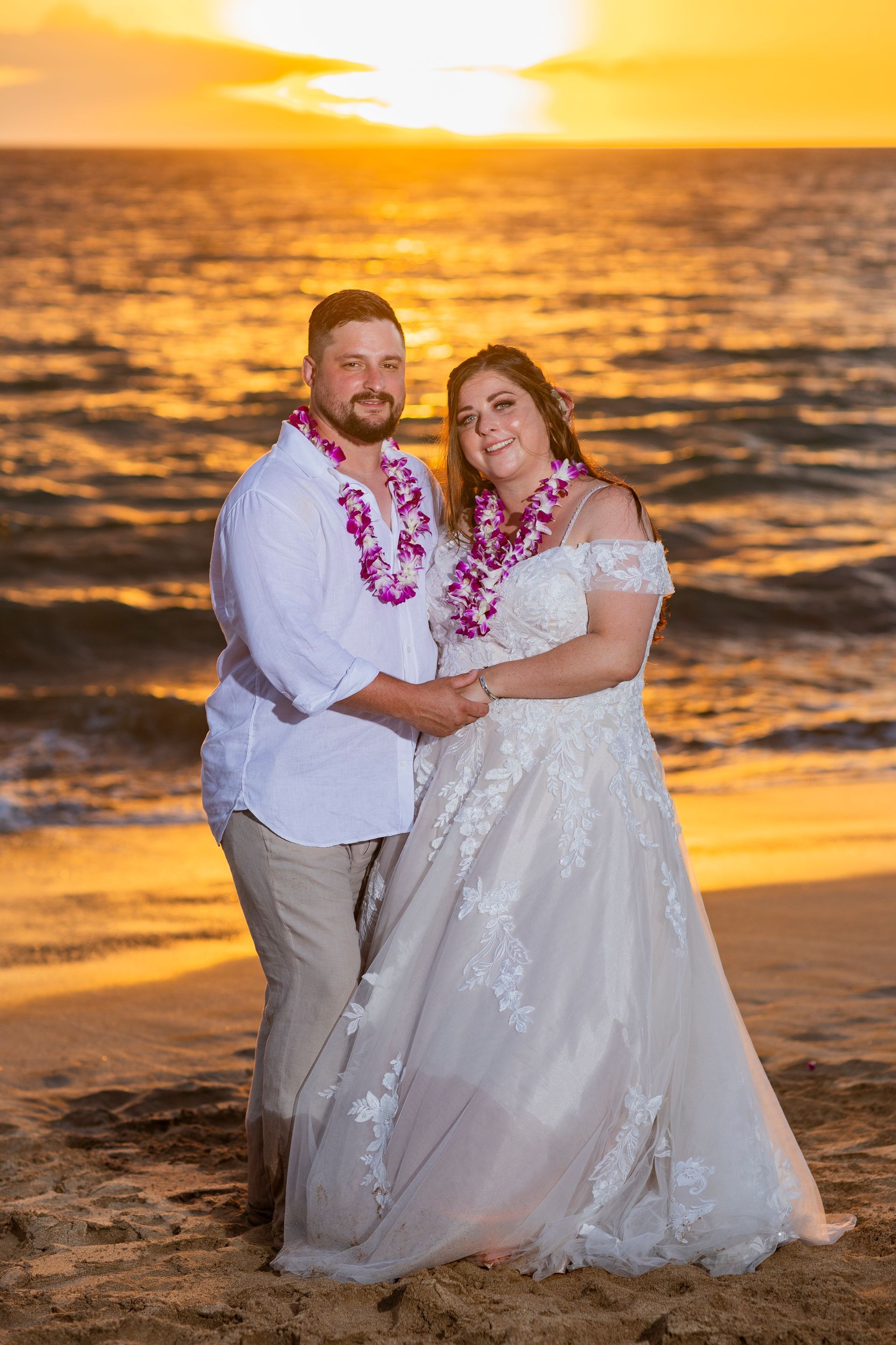 Couple on beach at sunset; man in white shirt, tan pants, woman in wedding dress, both wearing leis.