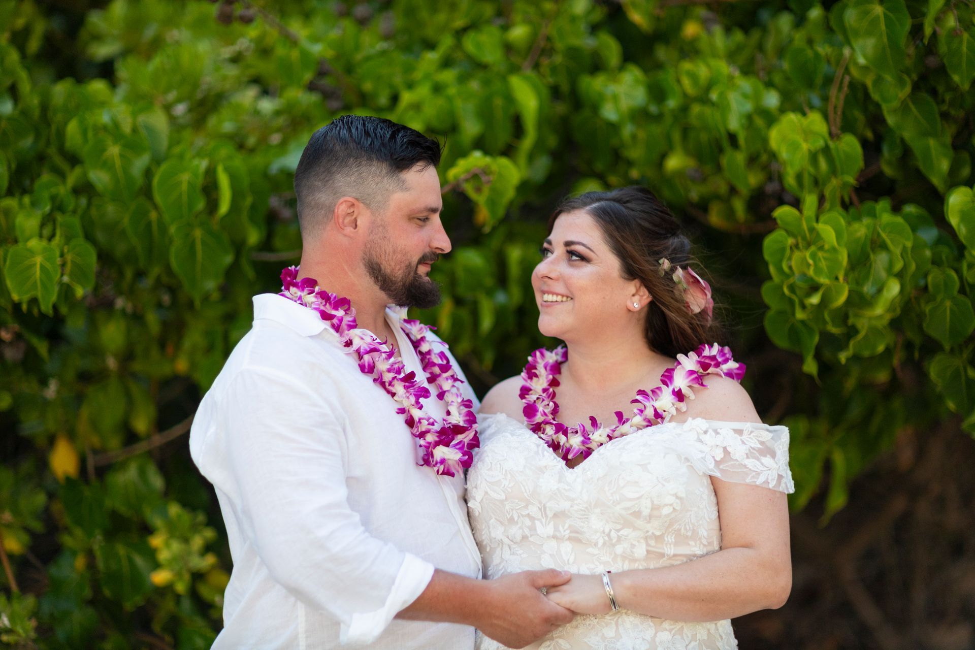 Couple in wedding attire, wearing leis, holding hands, smiling at each other, near green foliage.