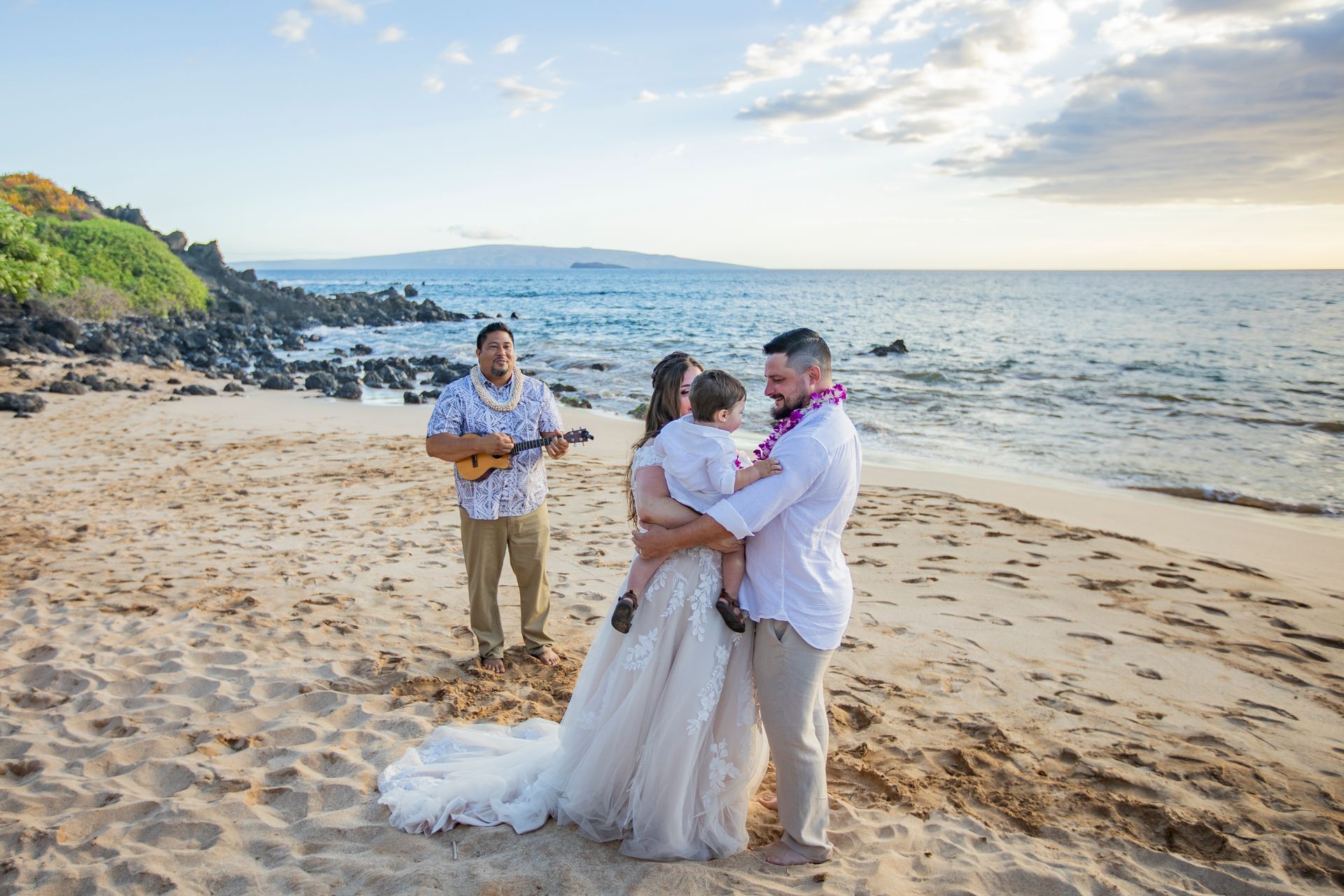 Wedding on a beach: couple embracing, musician plays ukulele, child in arms, ocean backdrop, sunny sky.