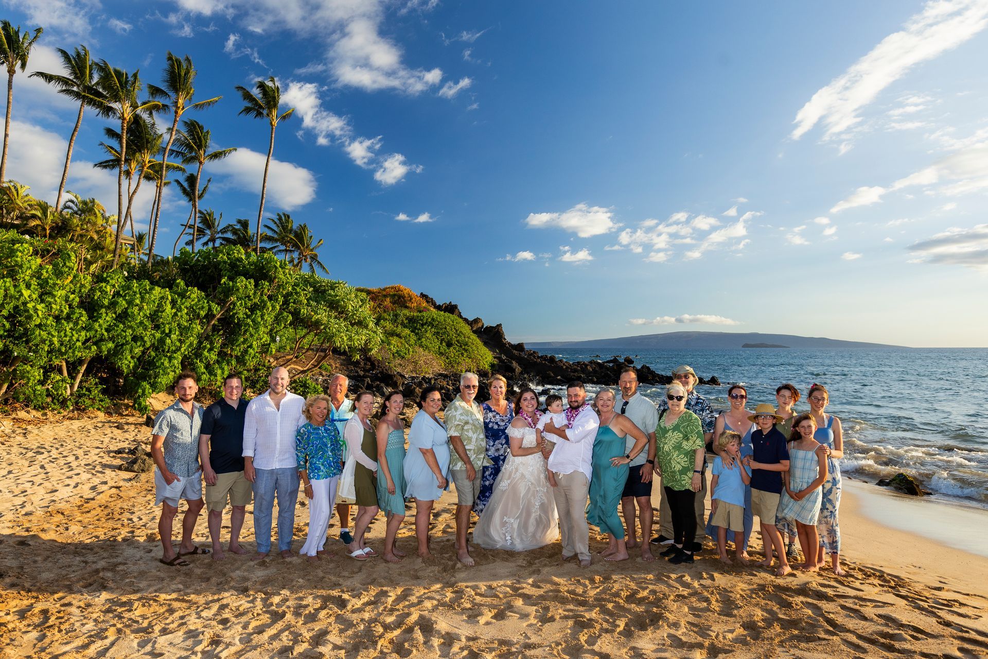 Large group of people, including a bride and groom, on a sunny beach with palm trees.