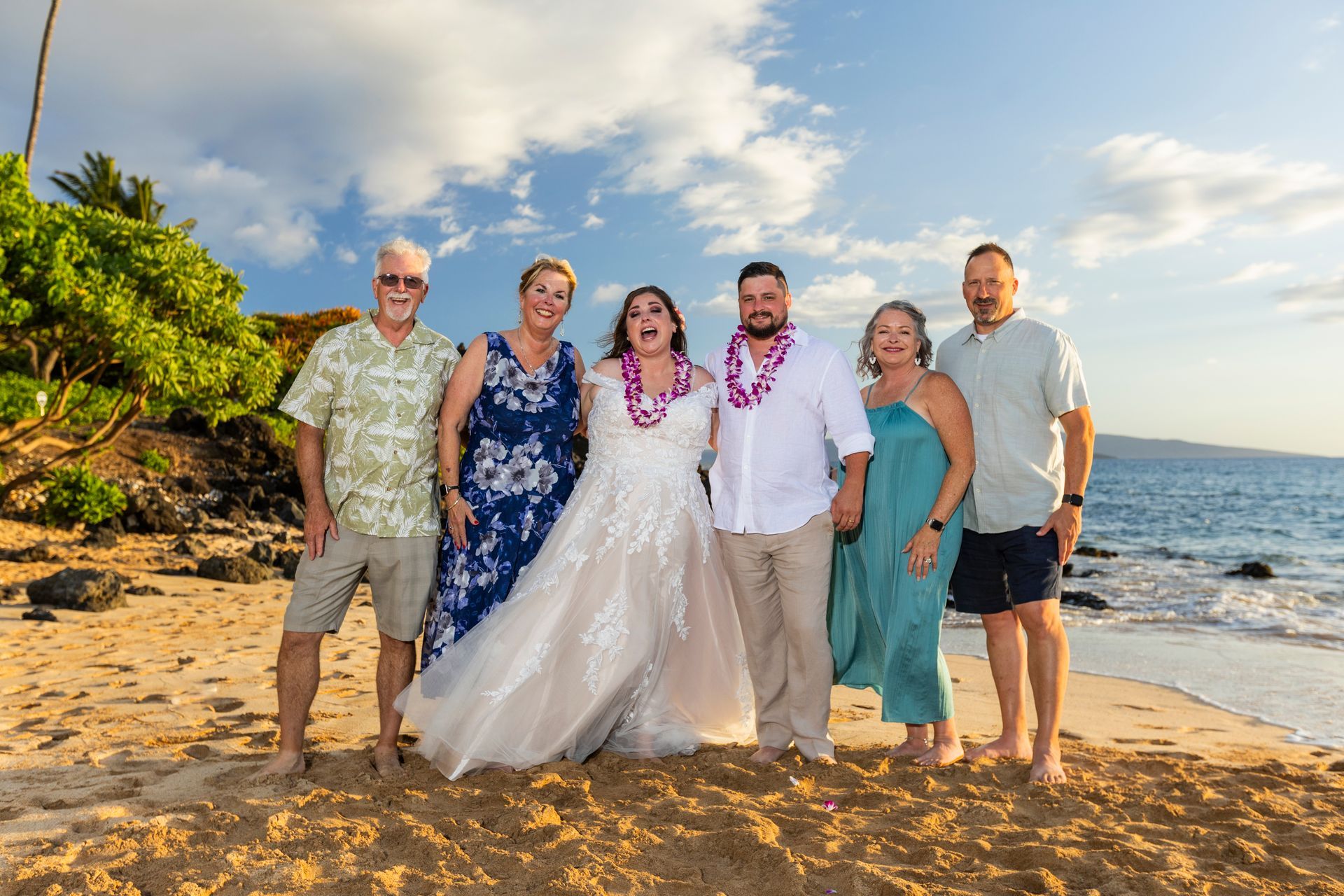 Wedding party poses on a beach: bride and groom flanked by parents under a partly cloudy sky.
